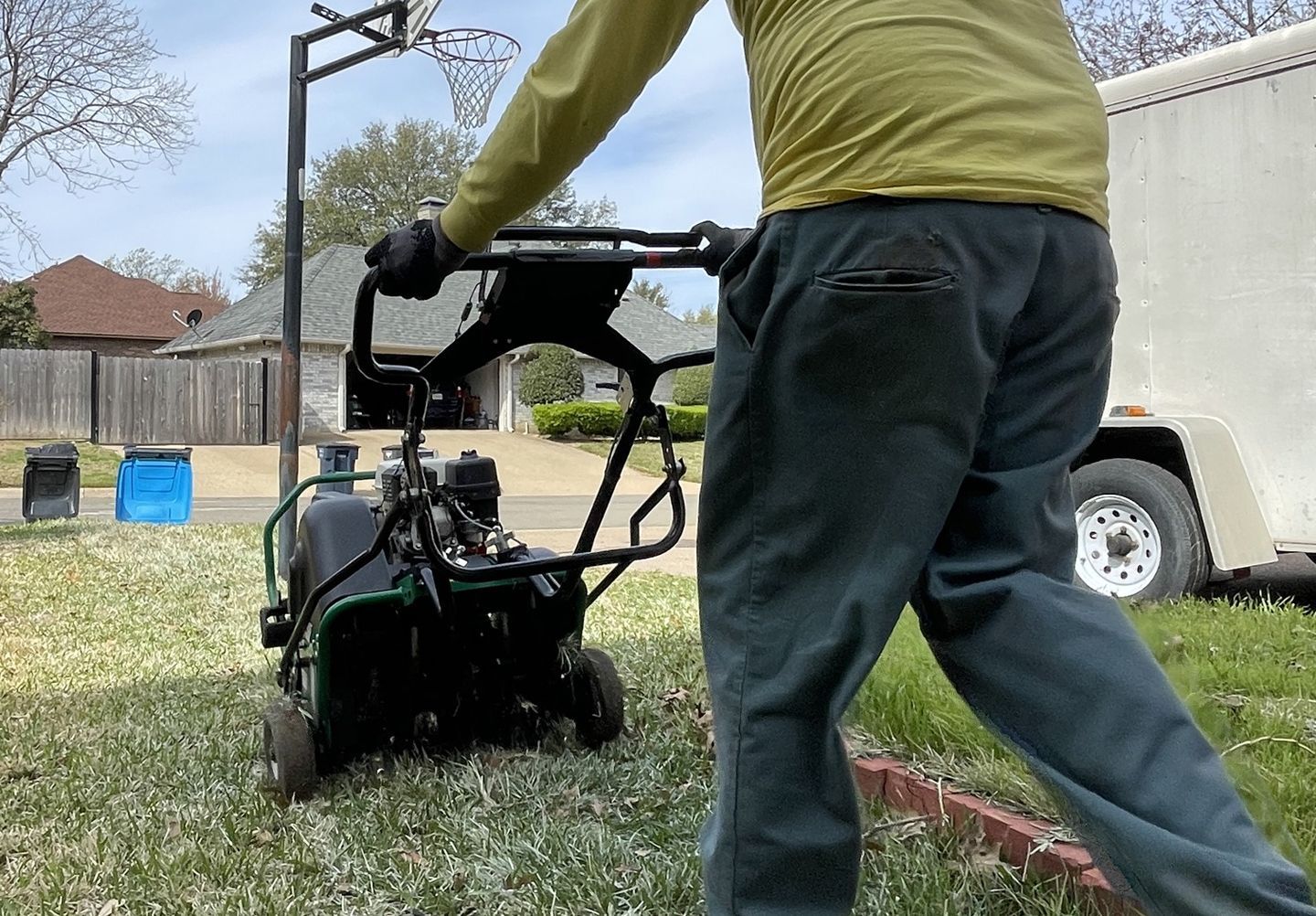 Person pushing a lawn aerator across a green lawn; a trailer is in the background.
