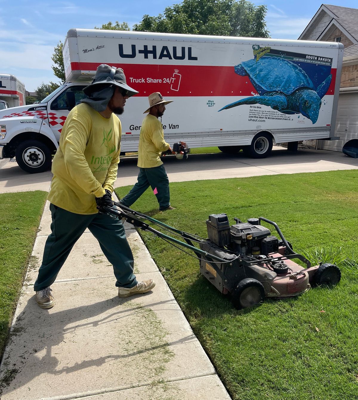 Two workers mowing grass beside a sidewalk with a lawnmower. U-Haul truck in background.