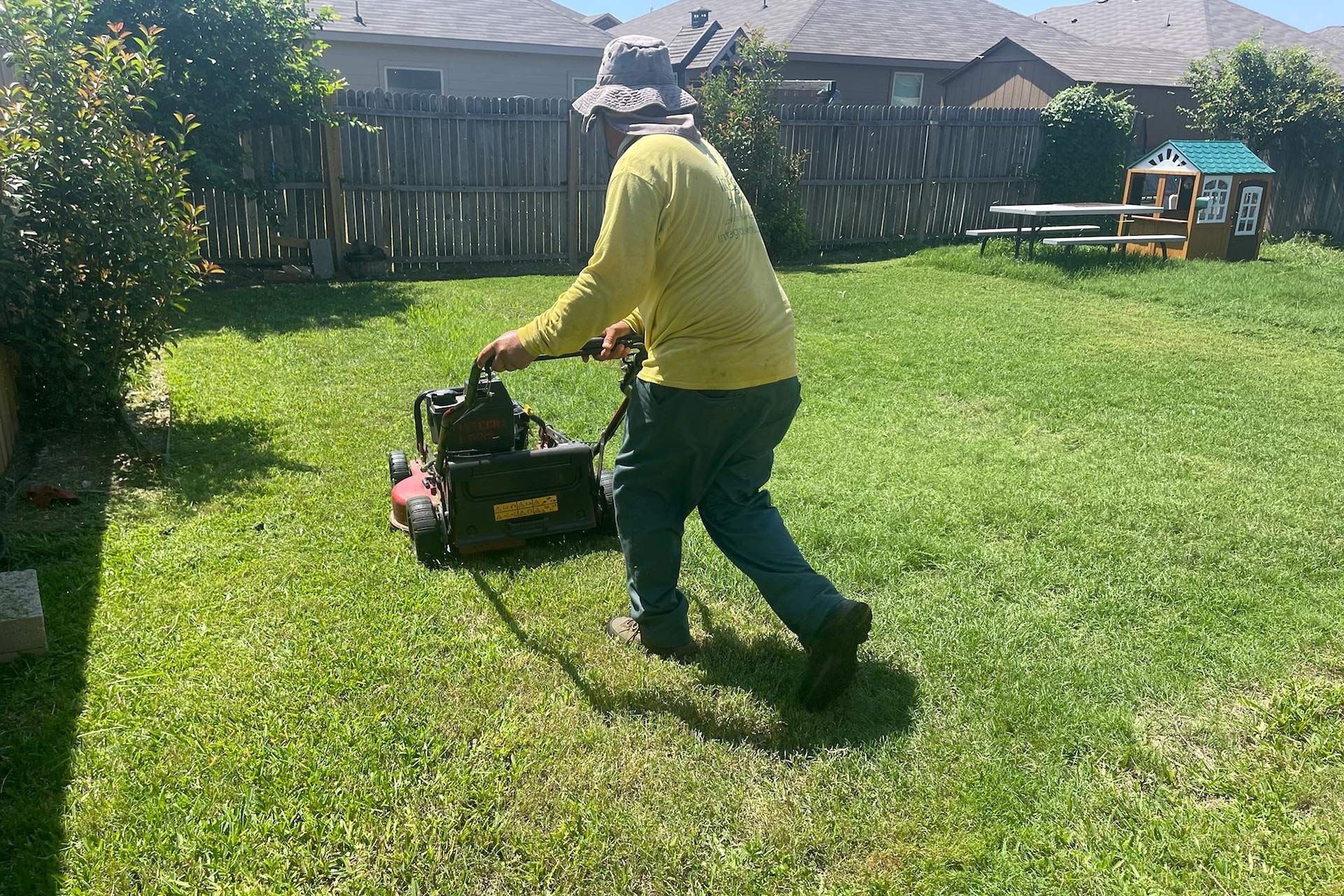 Person mowing a green lawn with a push mower in a backyard on a sunny day.