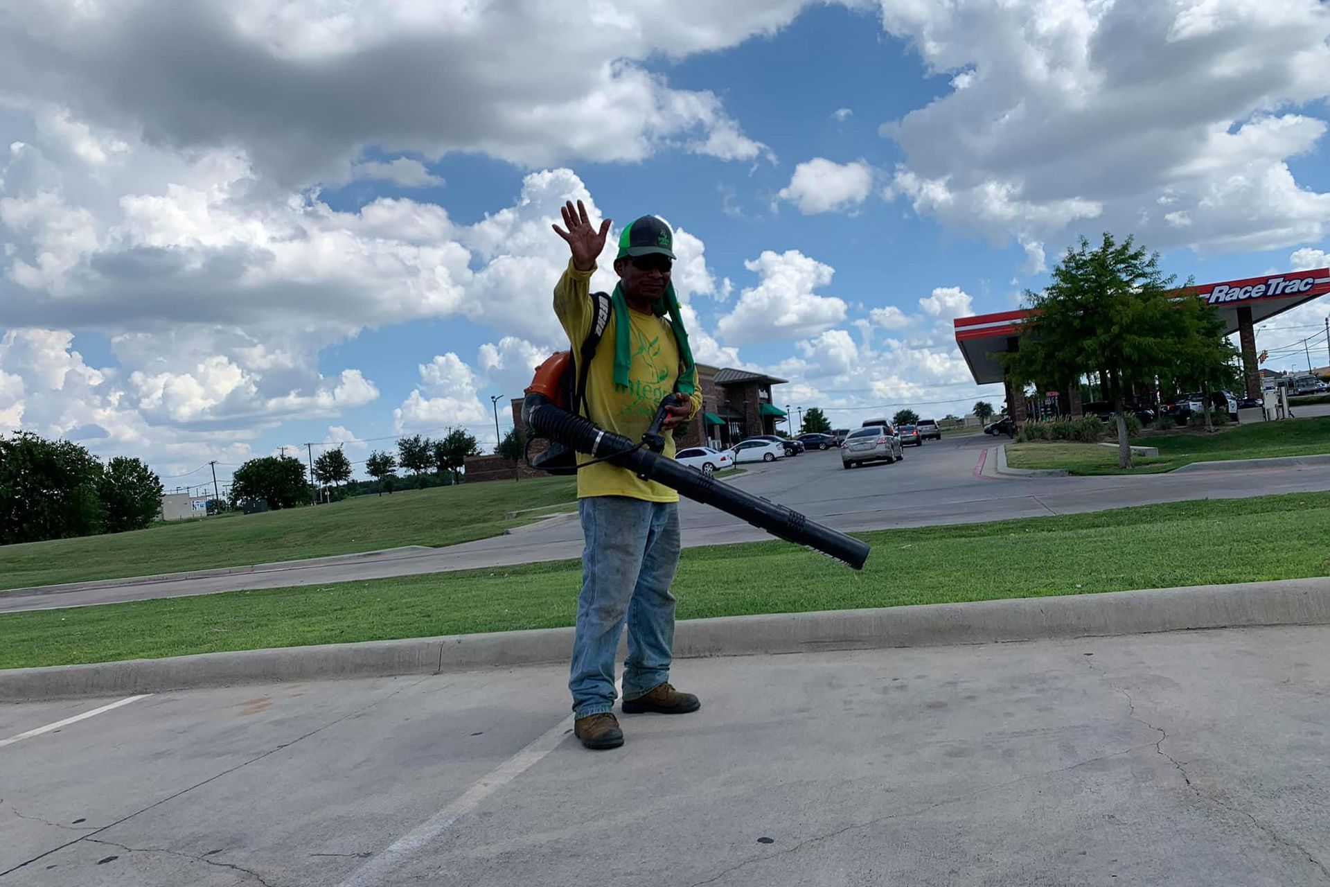 Man in work clothes waving, holding a leaf blower, standing on a parking lot, cloudy sky in background.