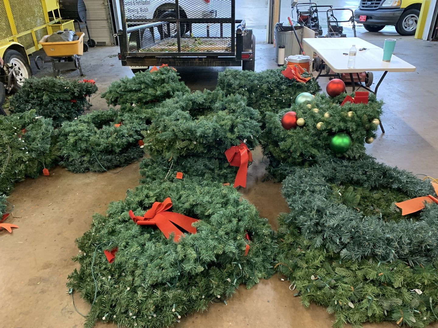 Wreaths with red bows and ornaments on floor, in a garage. A trailer and worktable are in the background.