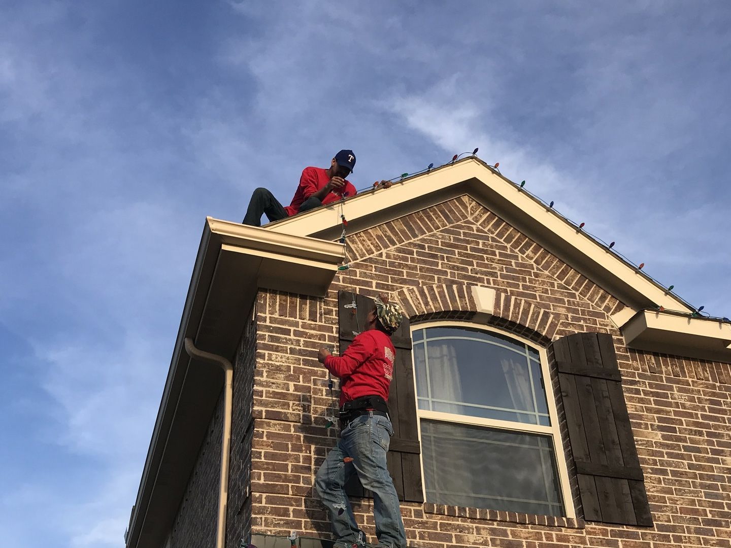 Two workers in red shirts installing Christmas lights on a brick house roof.