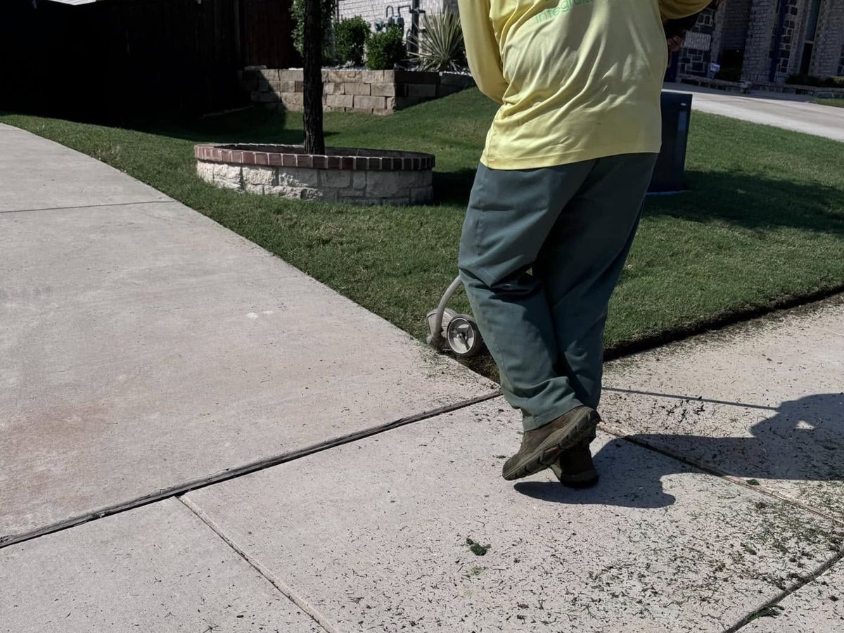 A person in yellow shirt edging the grass along a concrete sidewalk with a string trimmer.
