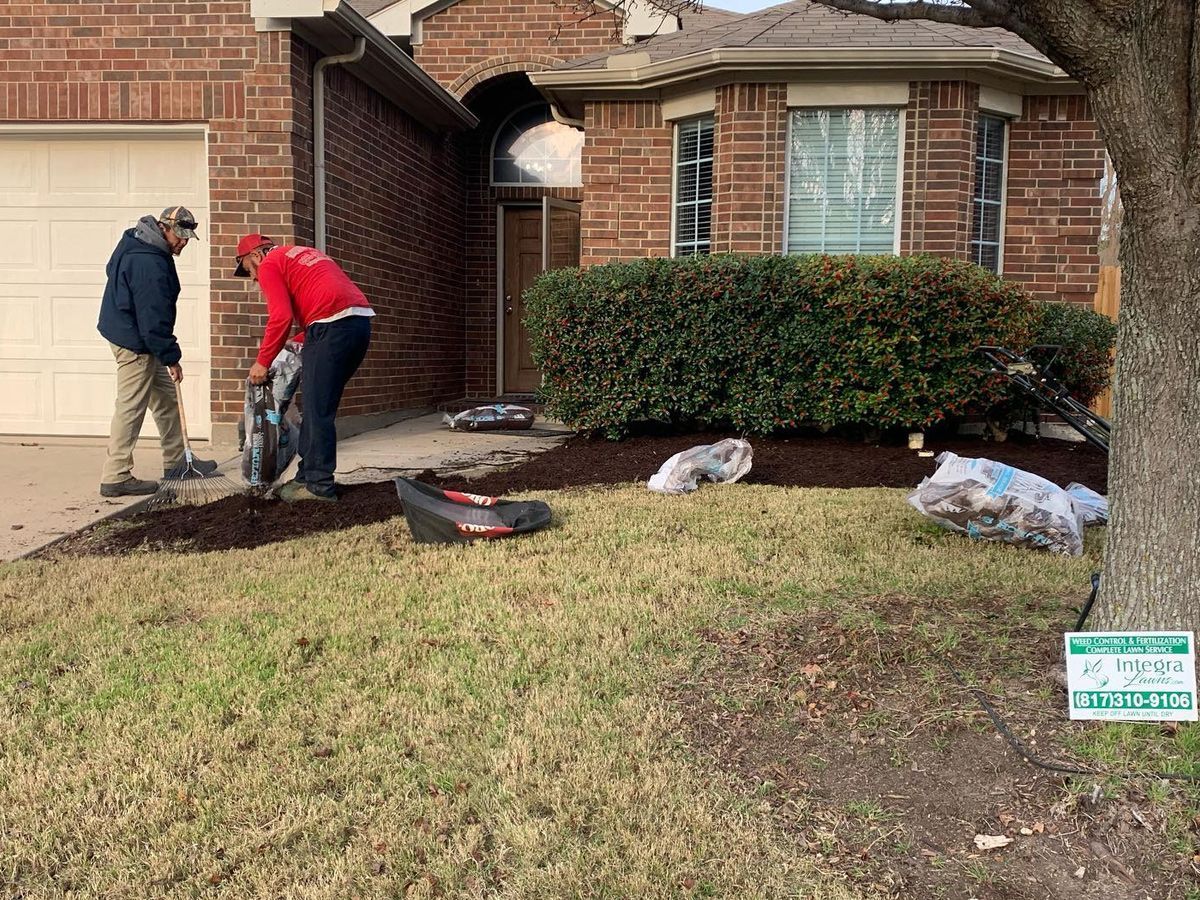 Two people spreading mulch in front yard of a brick house. Mulch bags and a lawnmower are visible.