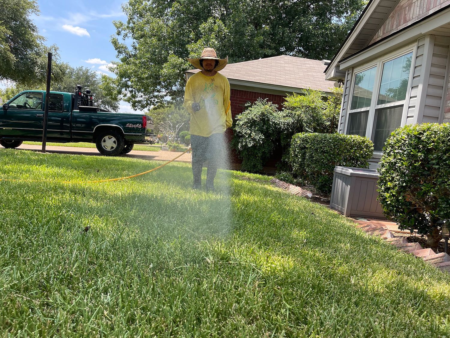 Person spraying lawn with green truck in background, house to the right. Bright sunlight.