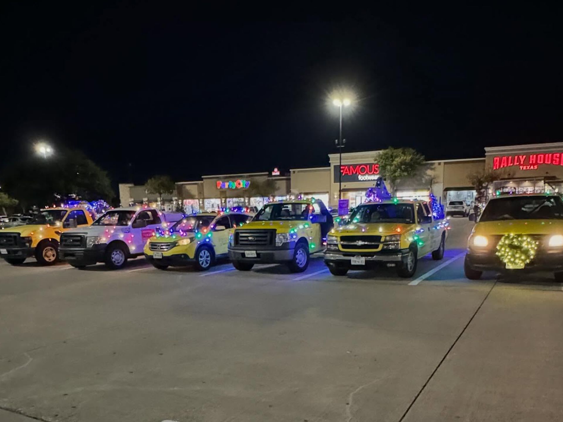 Vehicles decorated with colorful Christmas lights parked in a shopping center lot at night.