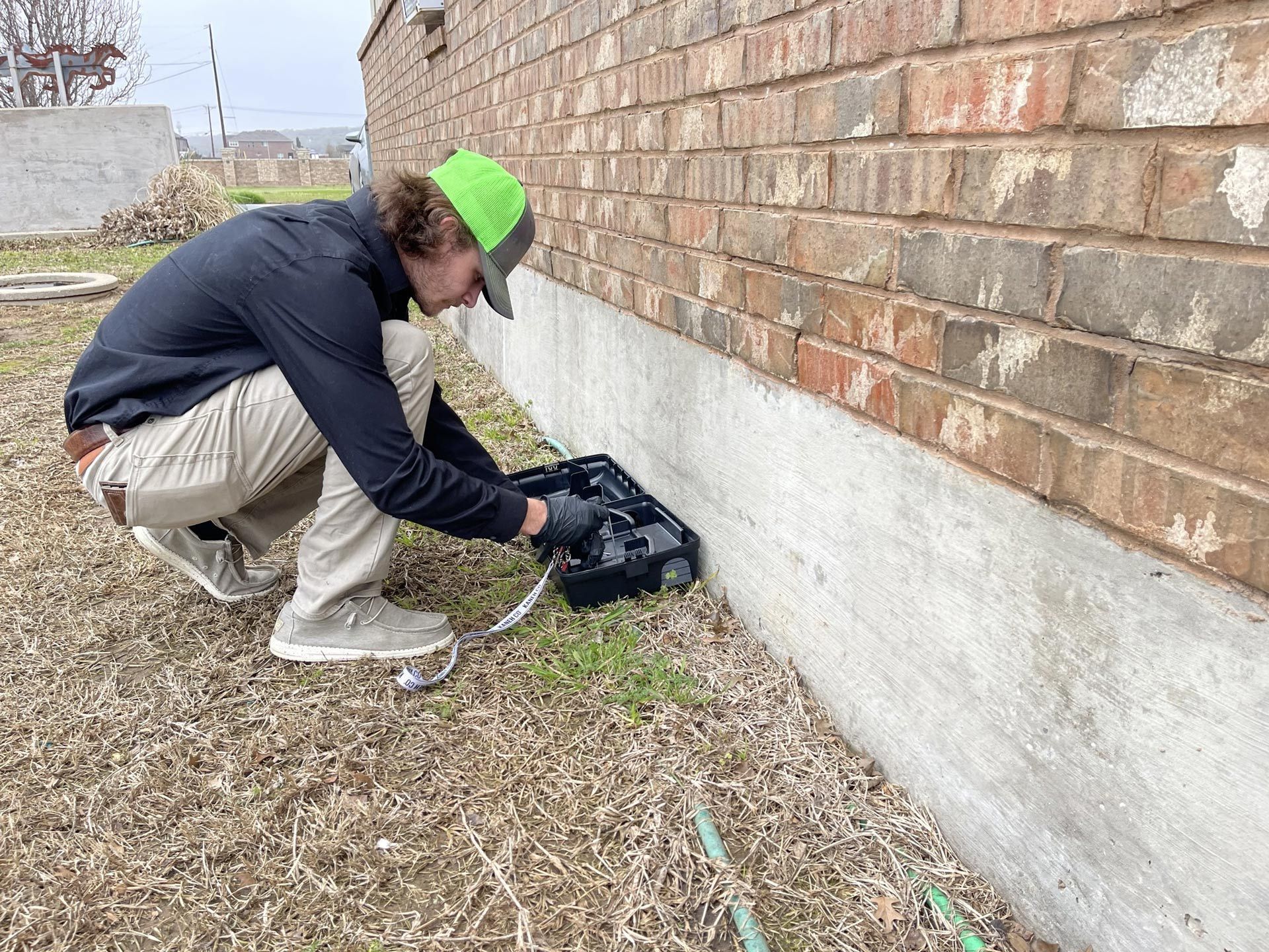 A person in a green cap crouches by a brick building, placing something in a black box.