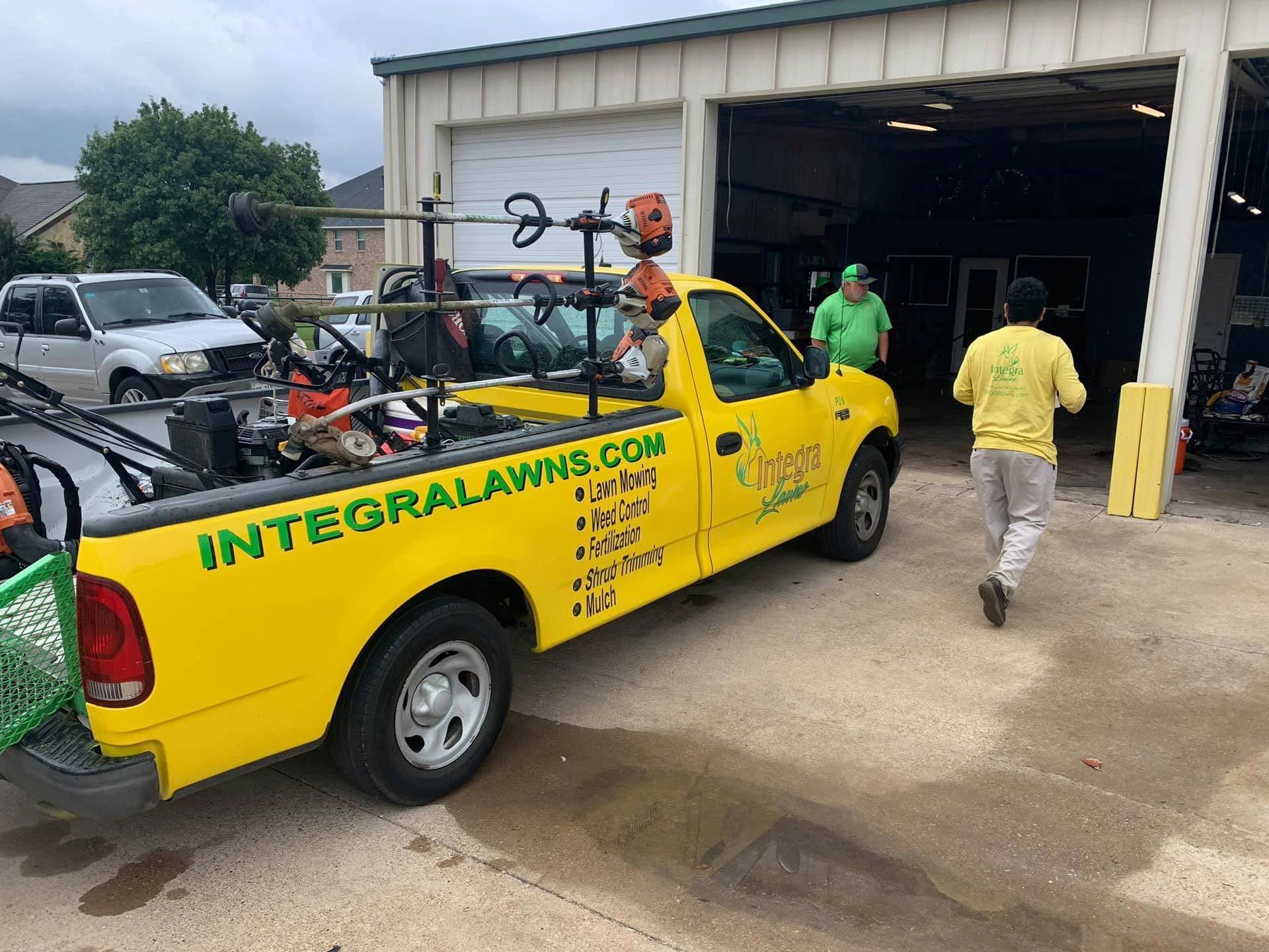 Yellow lawn care truck with equipment, two people near a garage. 