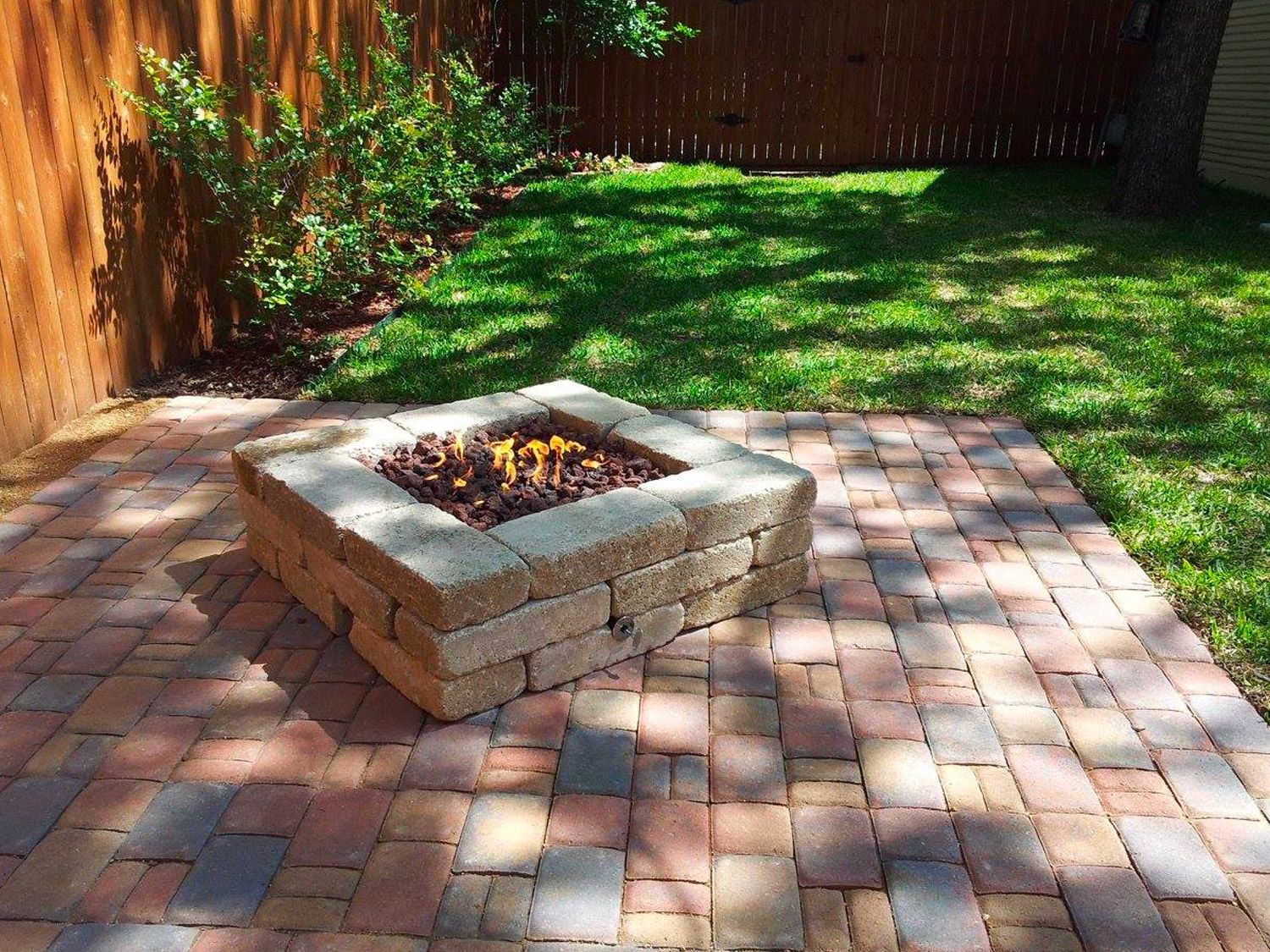 Stone fire pit on a brick patio in a backyard with green grass and a wooden fence.