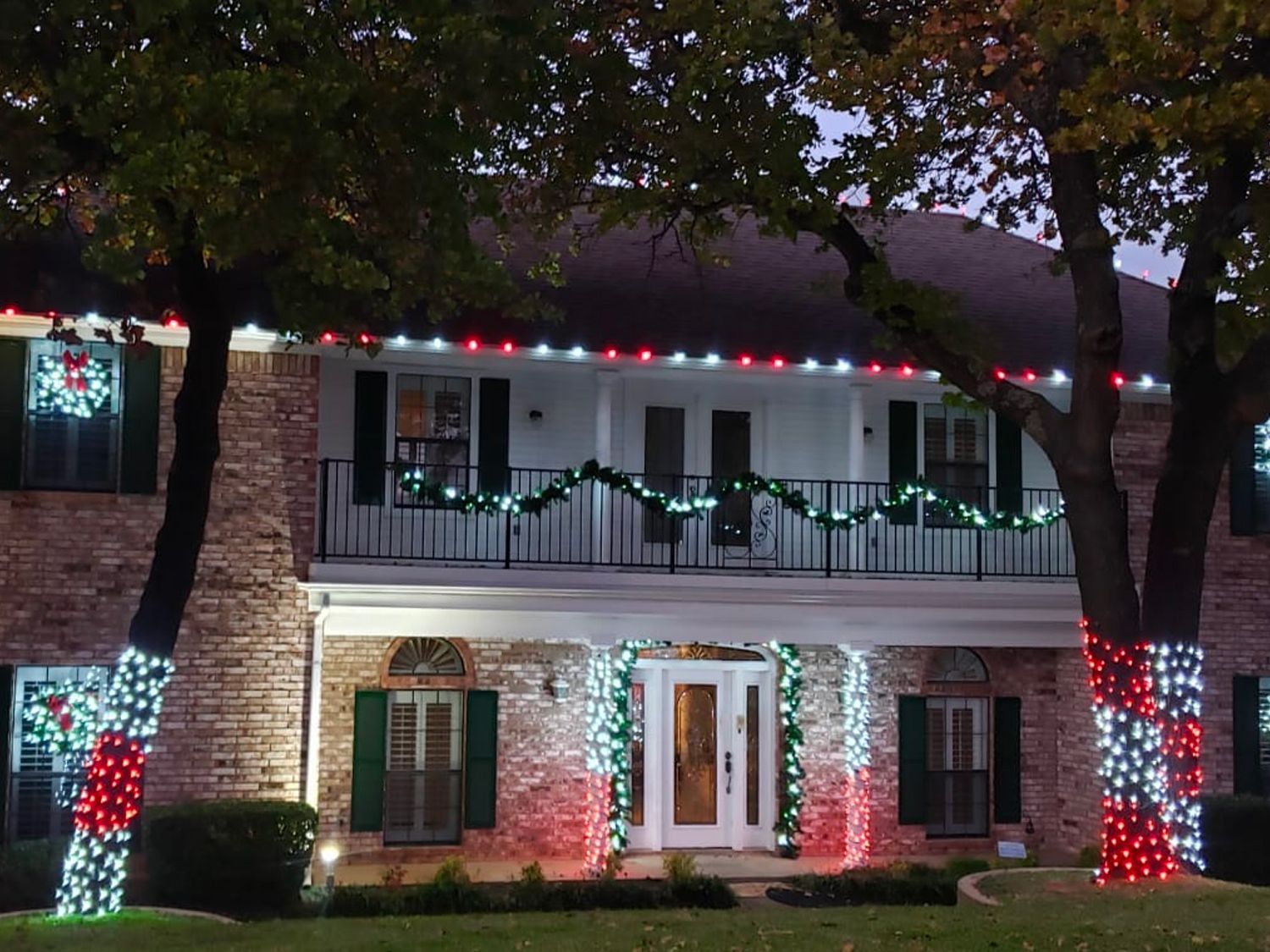 Two-story brick building decorated with Christmas lights: red, white, and green on trees, roofline, and around the door.