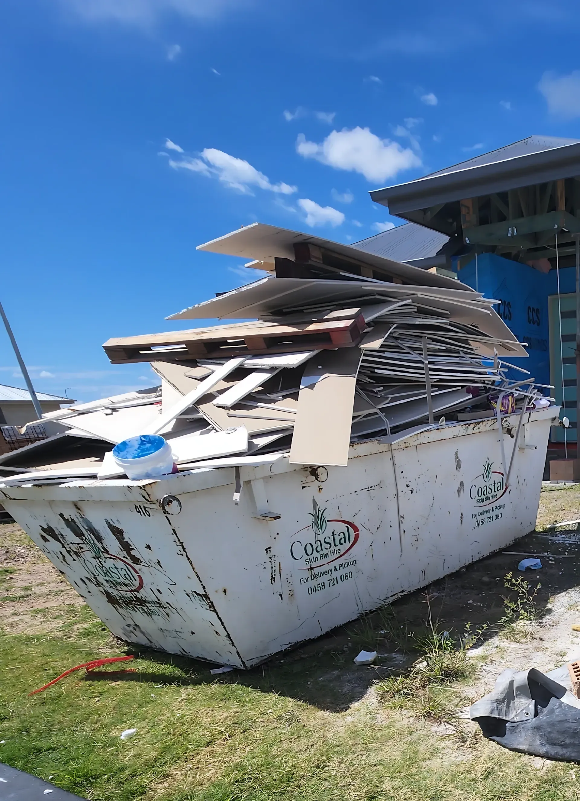 White dumpster overflowing with construction debris, in front of a building under construction, blue sky. — Coastal Skip Bin Hire In Maryborough, QLD