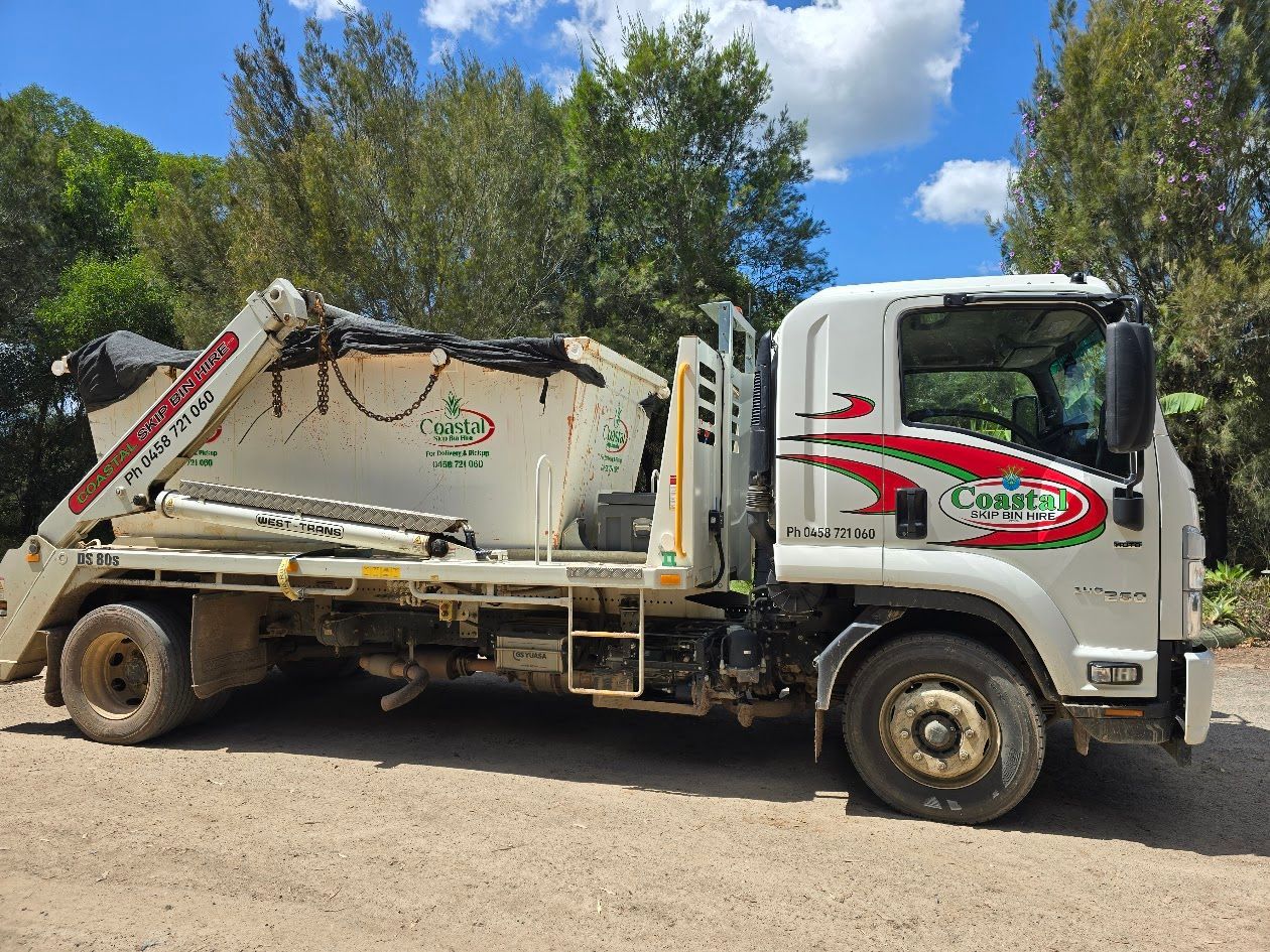 White waste removal truck with logo on the side, parked on a dirt road, trees in the background — Coastal Skip Bin Hire In Craignish, QLD