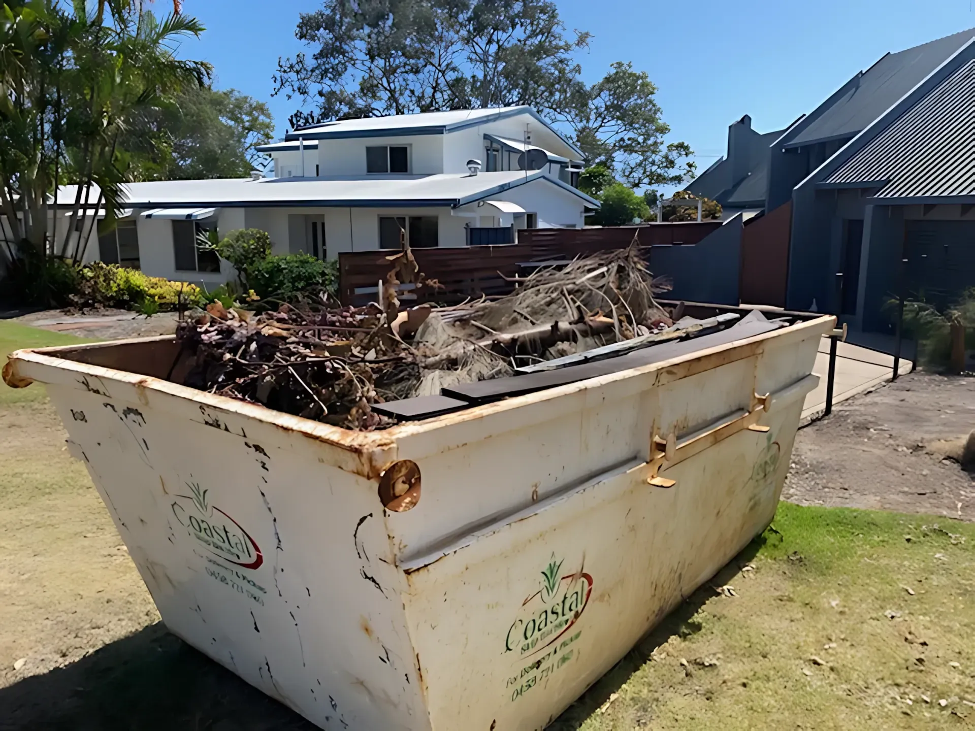 A large, overflowing dumpster with debris sits in front of two houses on a sunny day. — Coastal Skip Bin Hire In Urraween, QLD