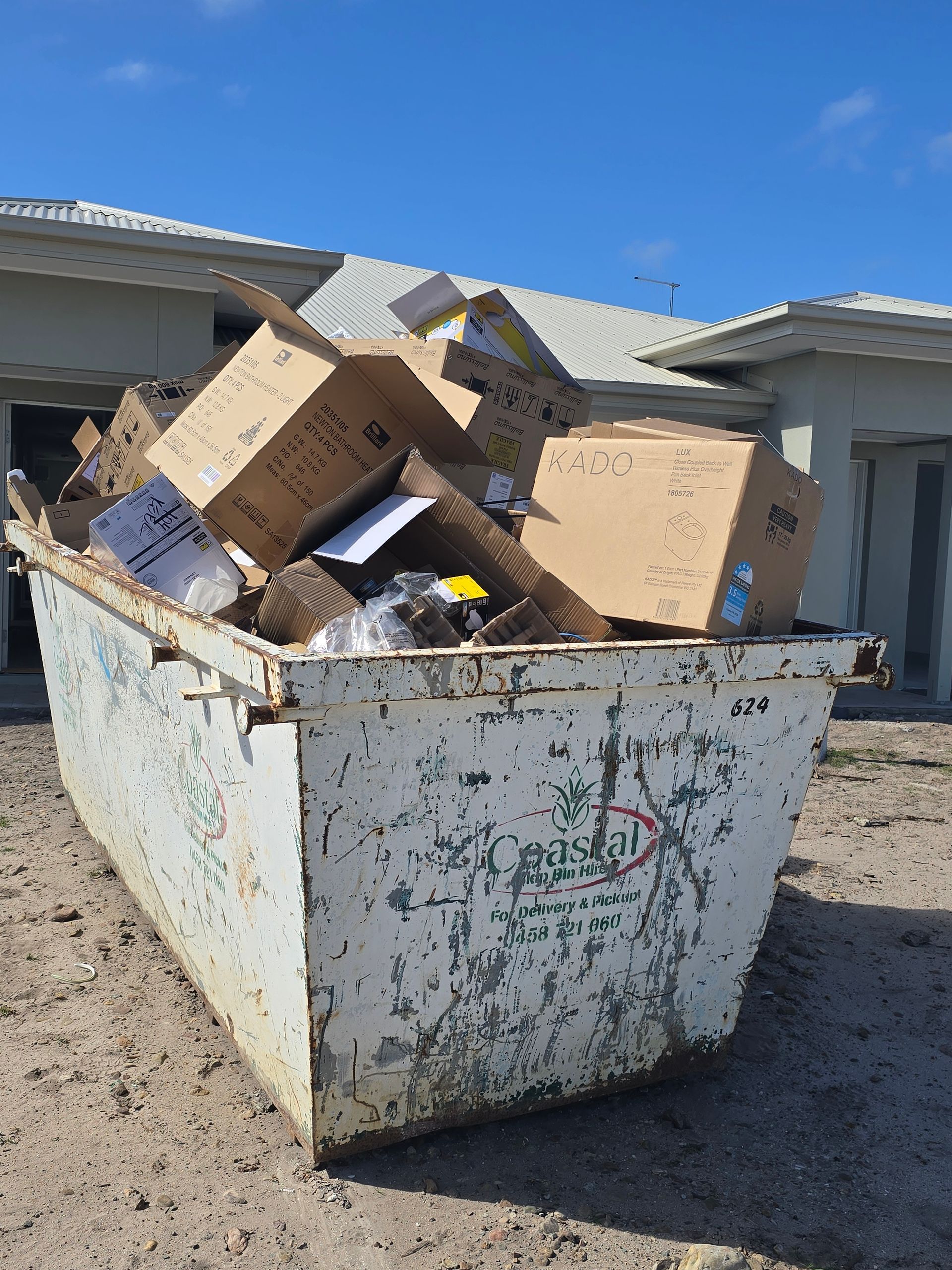 Large, overflowing dumpster filled with cardboard boxes, in front of a light-colored house under a blue sky — Coastal Skip Bin Hire In Craignish, QLD