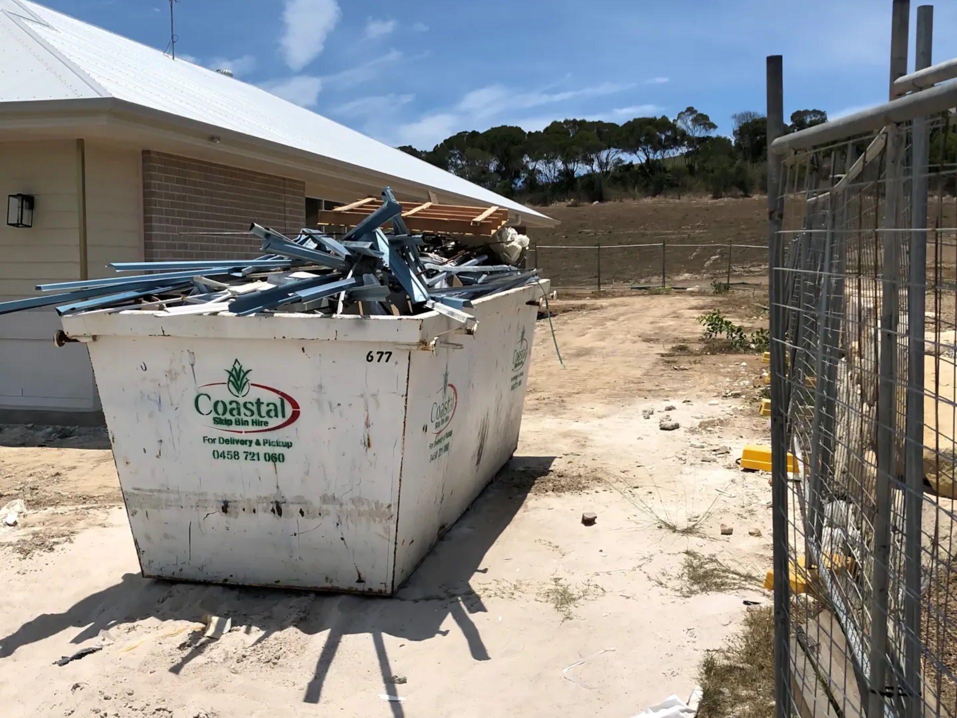 White dumpster filled with debris on a construction site next to a building and fence on a sunny day — Coastal Skip Bin Hire In Craignish, QLD