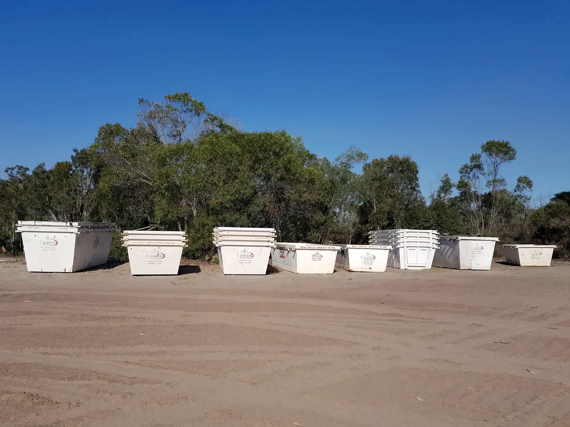 White skip bins in a row on a dirt lot, trees and blue sky in the background. — Coastal Skip Bin Hire In Craignish, QLD