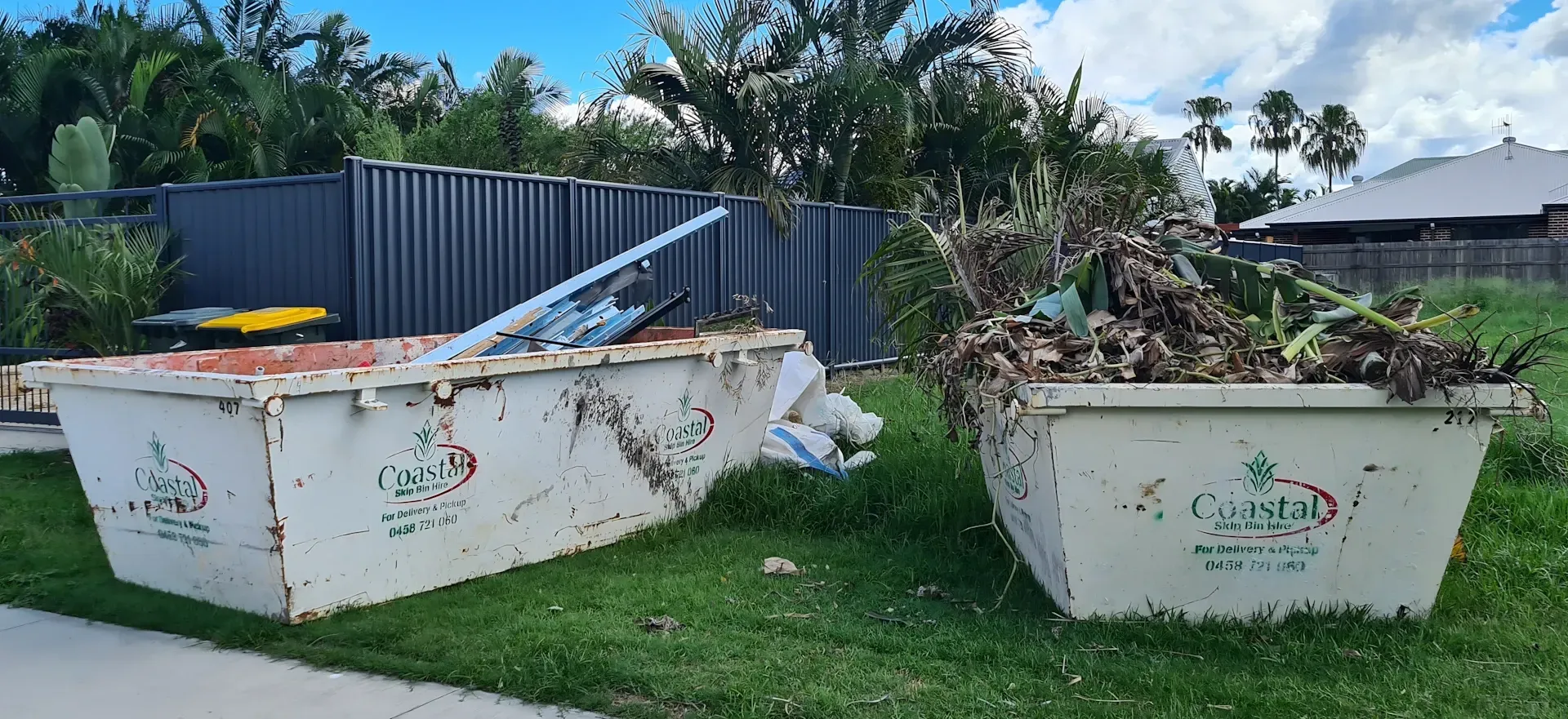 Two white dumpsters on grass, filled with trash, in front of a fence and houses. — Coastal Skip Bin Hire In Fraser Coast, QLD