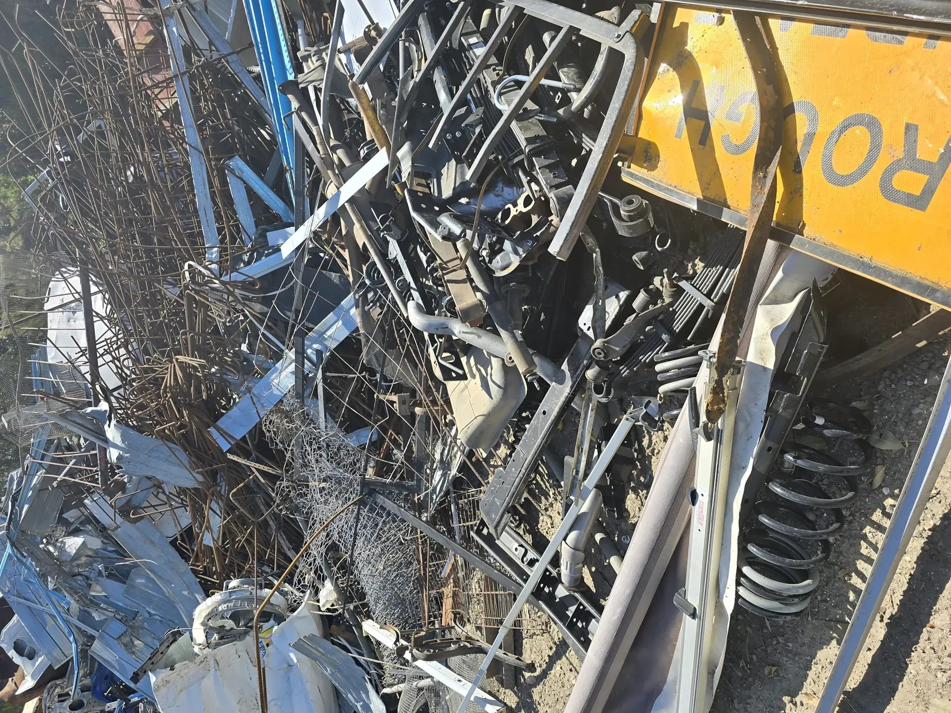 Pile of mixed scrap metal in sunlight, including wires, springs, and car parts; yellow sign visible. — Coastal Skip Bin Hire In Eli Waters, QLD