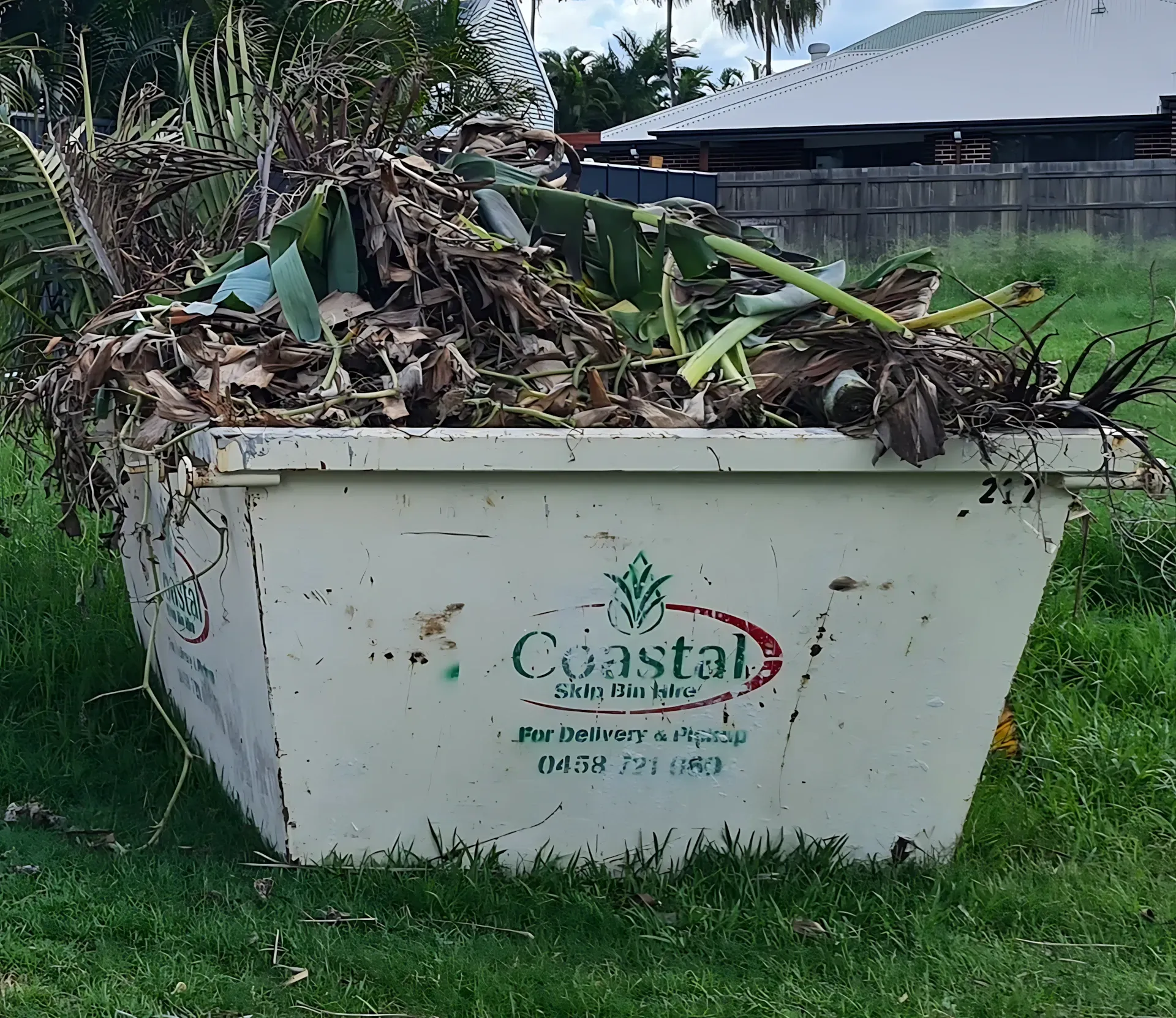 A white Coastal Skips dumpster overflowing with green and brown yard waste, sitting on grass. — Coastal Skip Bin Hire In Maryborough, QLD