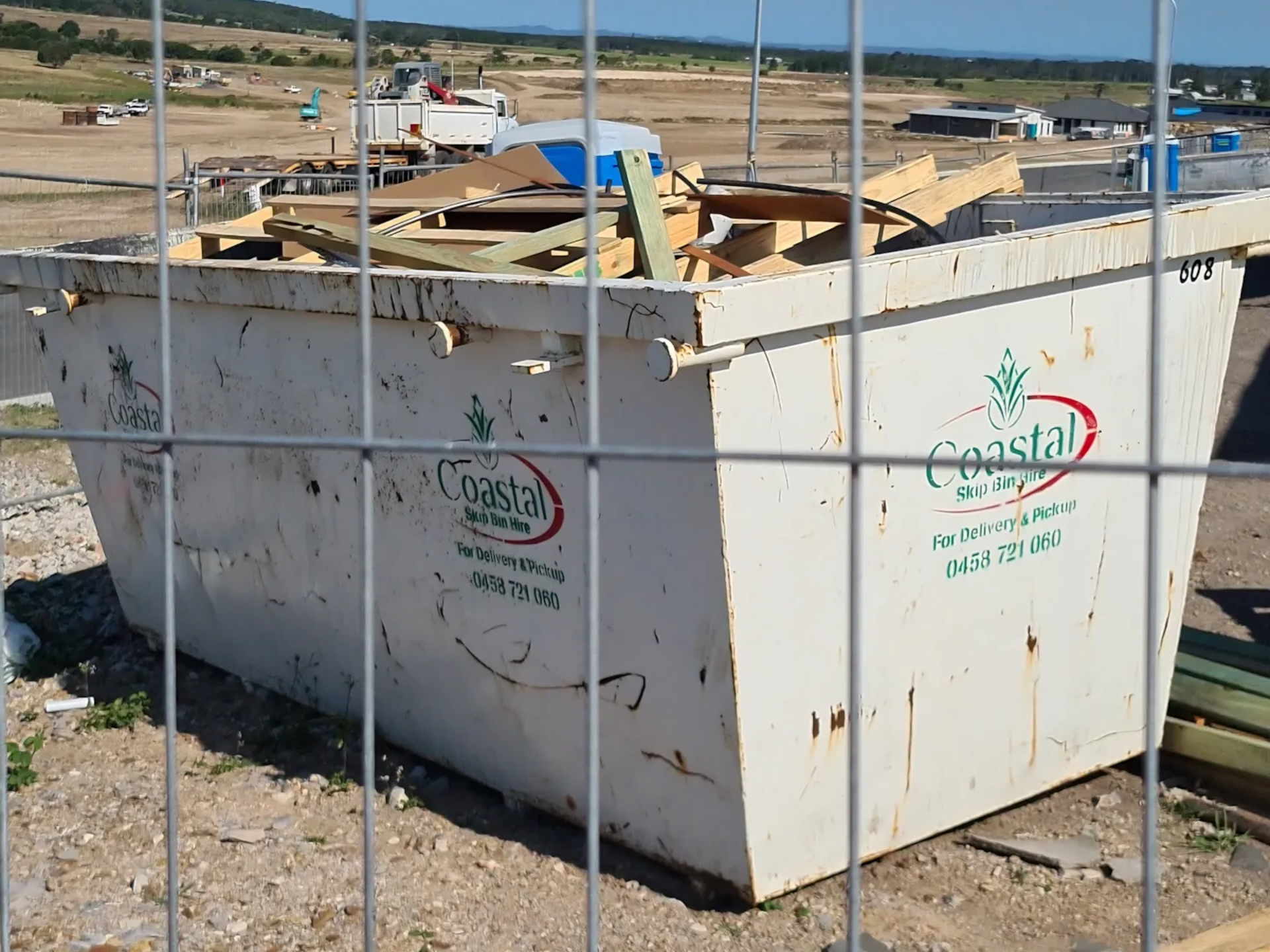 Metal dumpster filled with construction debris, behind a wire fence at a construction site. 