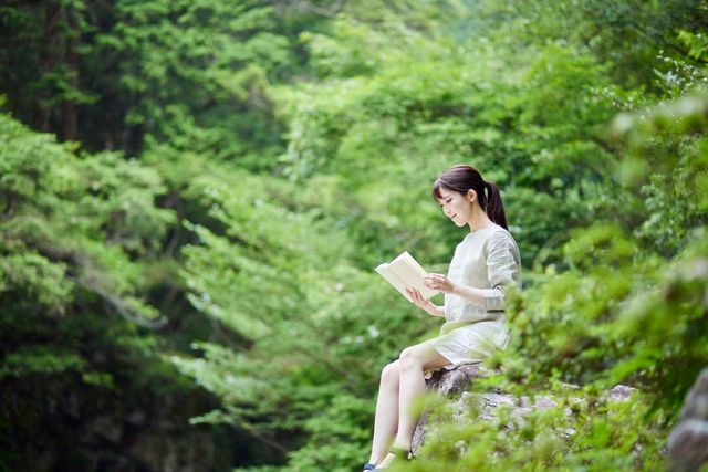 A woman is sitting on a rock reading a book in the woods.