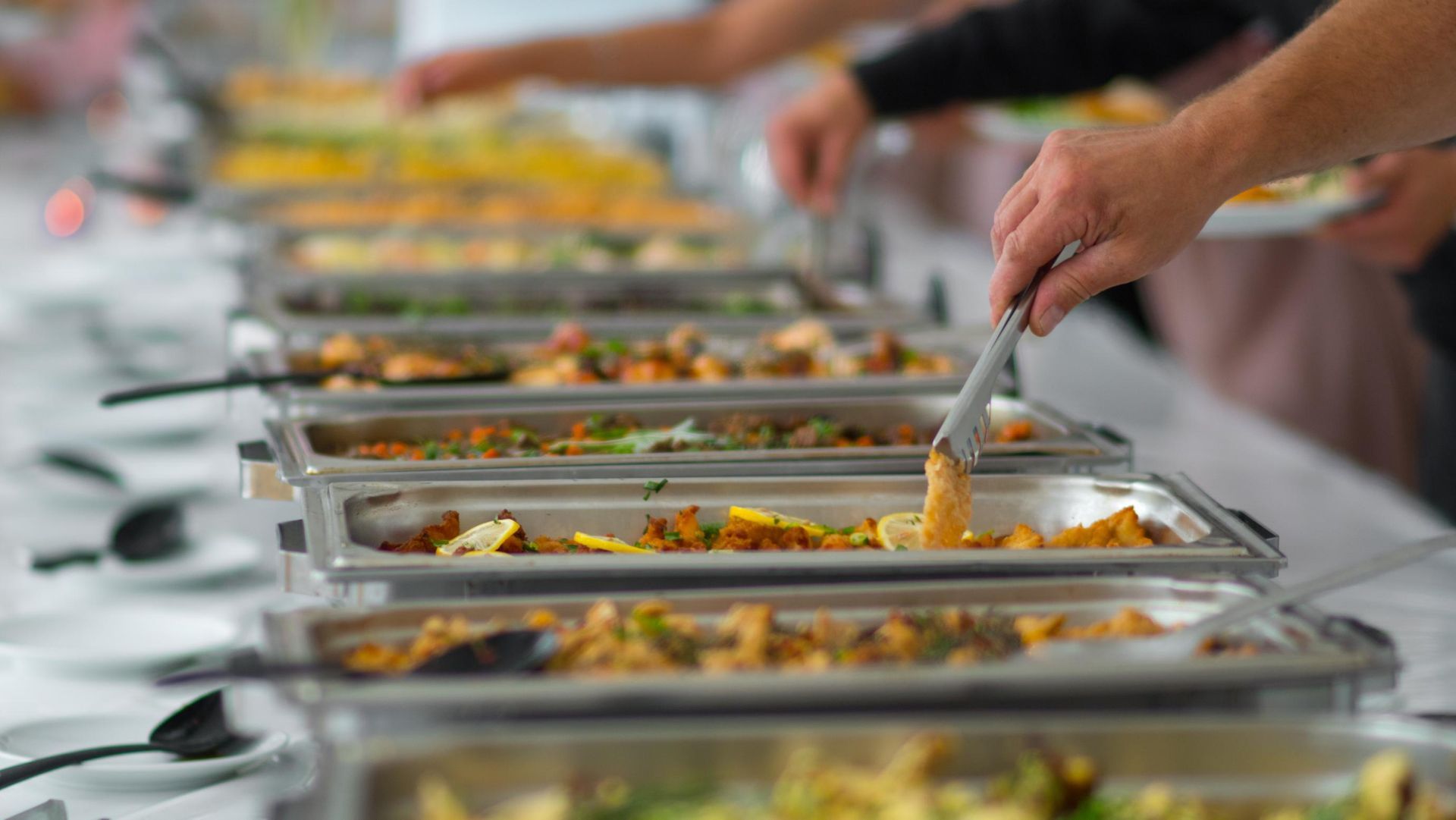 People serving food from stainless steel buffet trays with tongs at a catered event