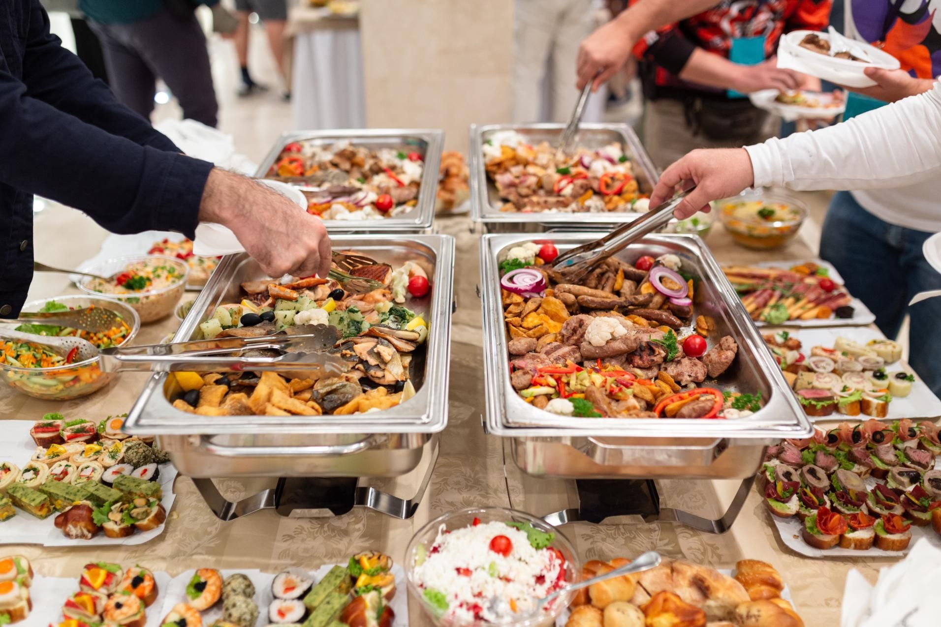 Buffet table with trays of assorted dishes and people serving themselves in a bright dining hall