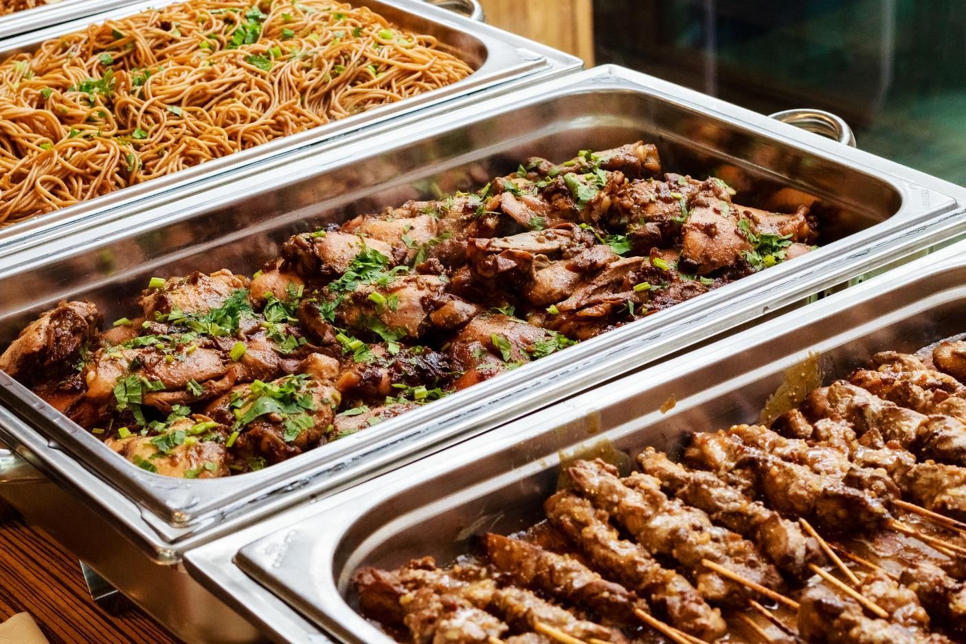 Three buffet trays of noodles, grilled meat, and skewered food on a serving table