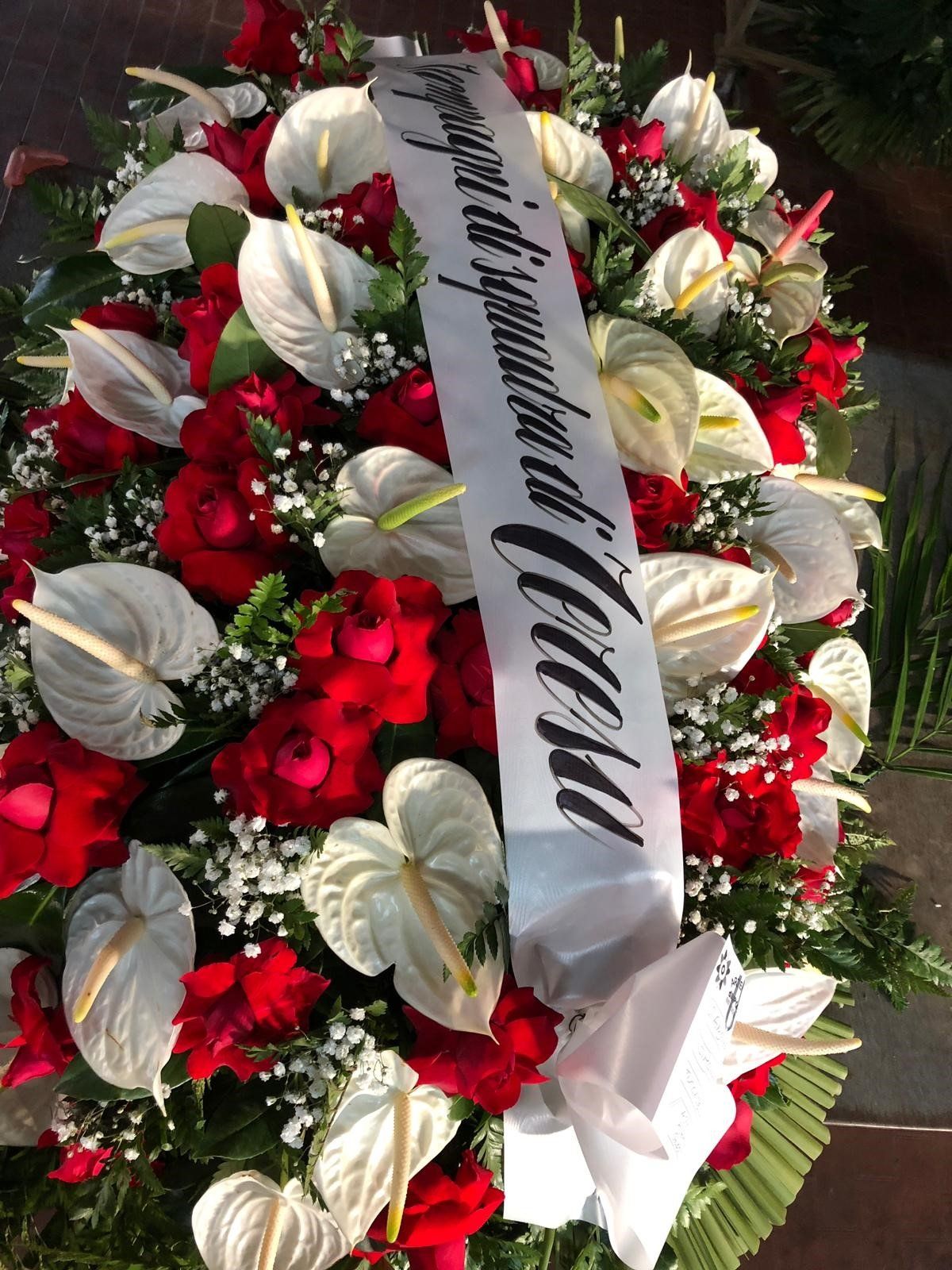 Corona funebre con rose rosse, anthurium bianchi e un nastro bianco.