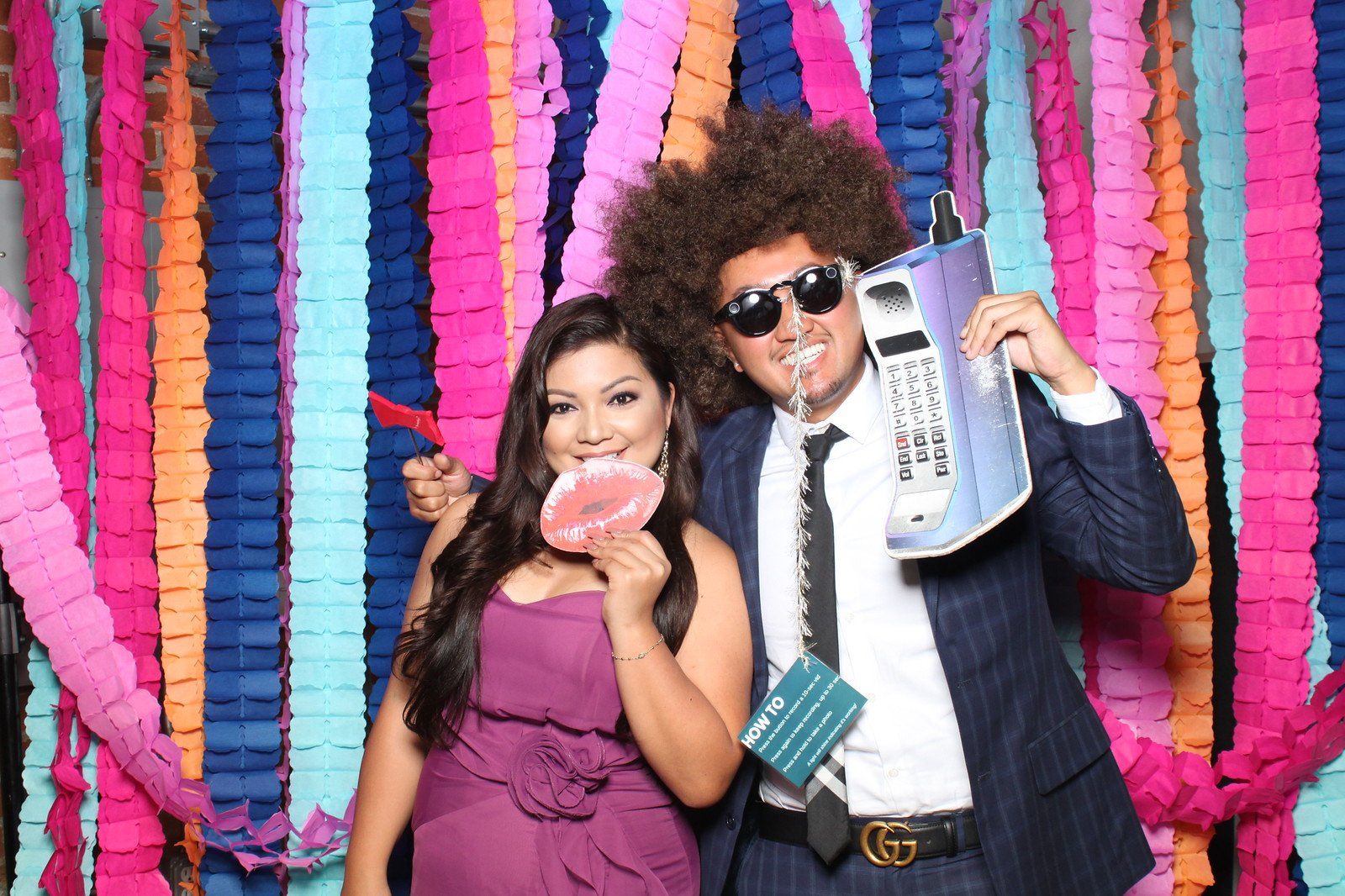 Woman in purple dress and man in afro, suit, and sunglasses pose with props in front of colorful streamers.