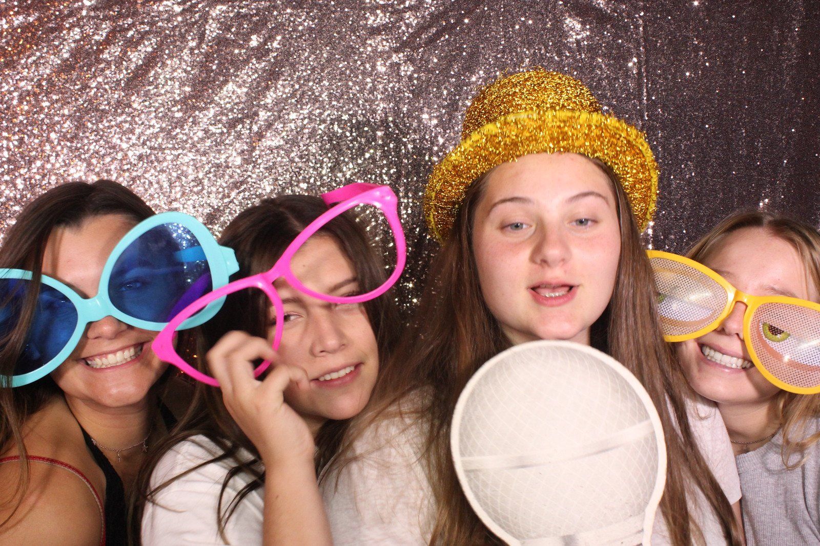 Four people in front of a shiny backdrop, wearing oversized novelty glasses and a gold hat, around a microphone.