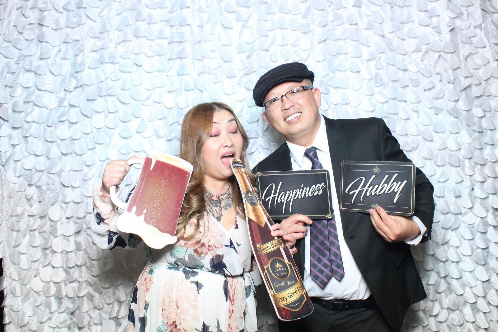 Couple in photo booth holding props; woman drinking from champagne bottle cutout, man holding