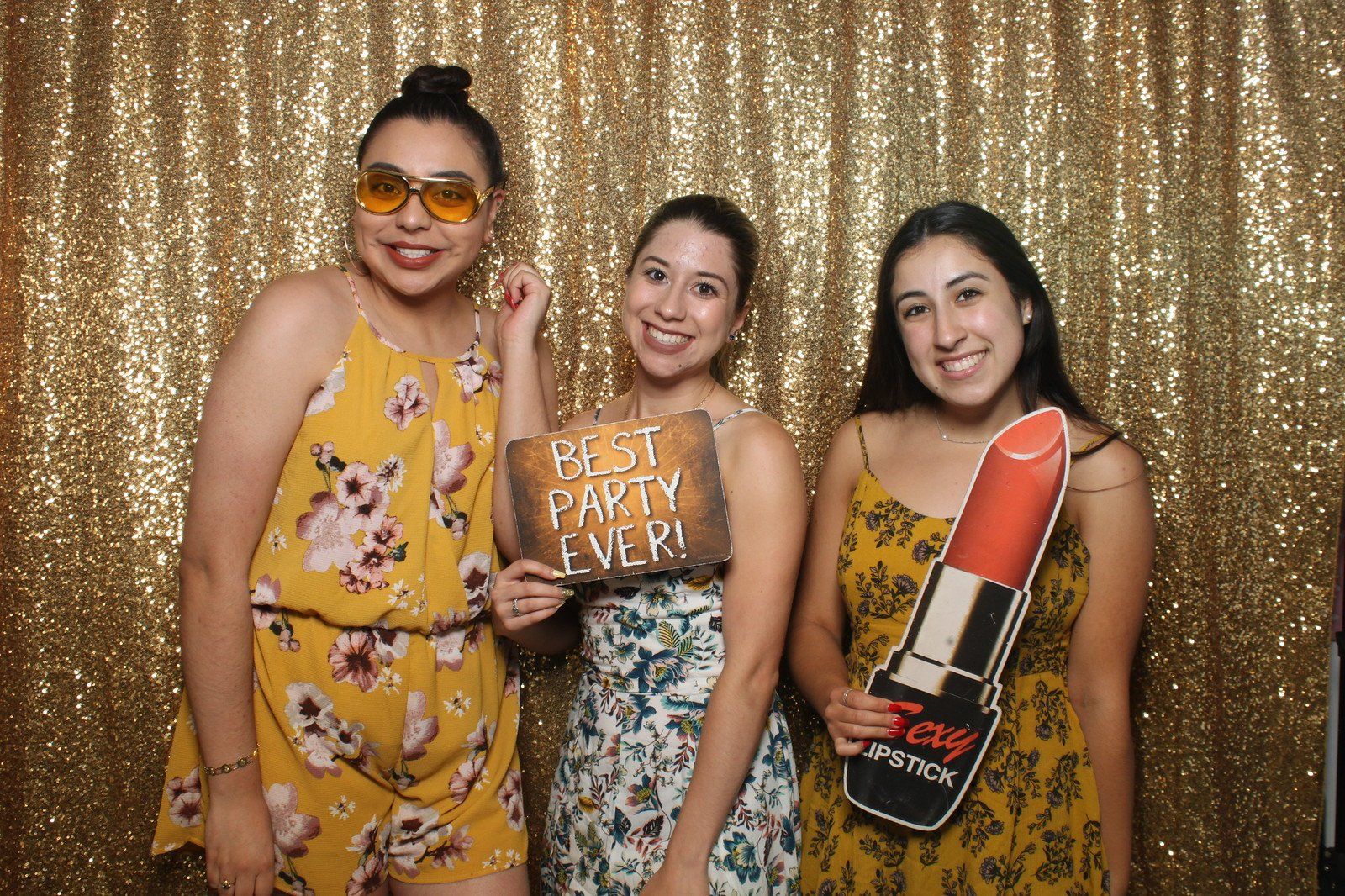 Three smiling people posing in front of a gold sequin backdrop, holding photo booth props.
