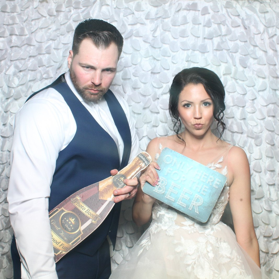 Man in vest holds champagne prop, woman in wedding dress puckers; photo booth.