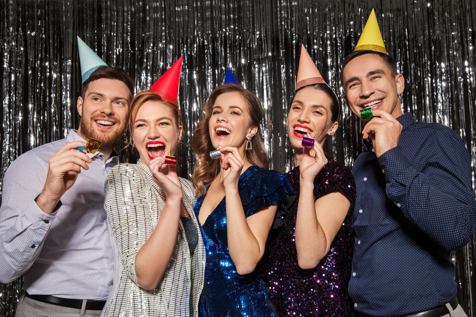 Five people with party hats, blowing noisemakers, smiling in front of a silver curtain.