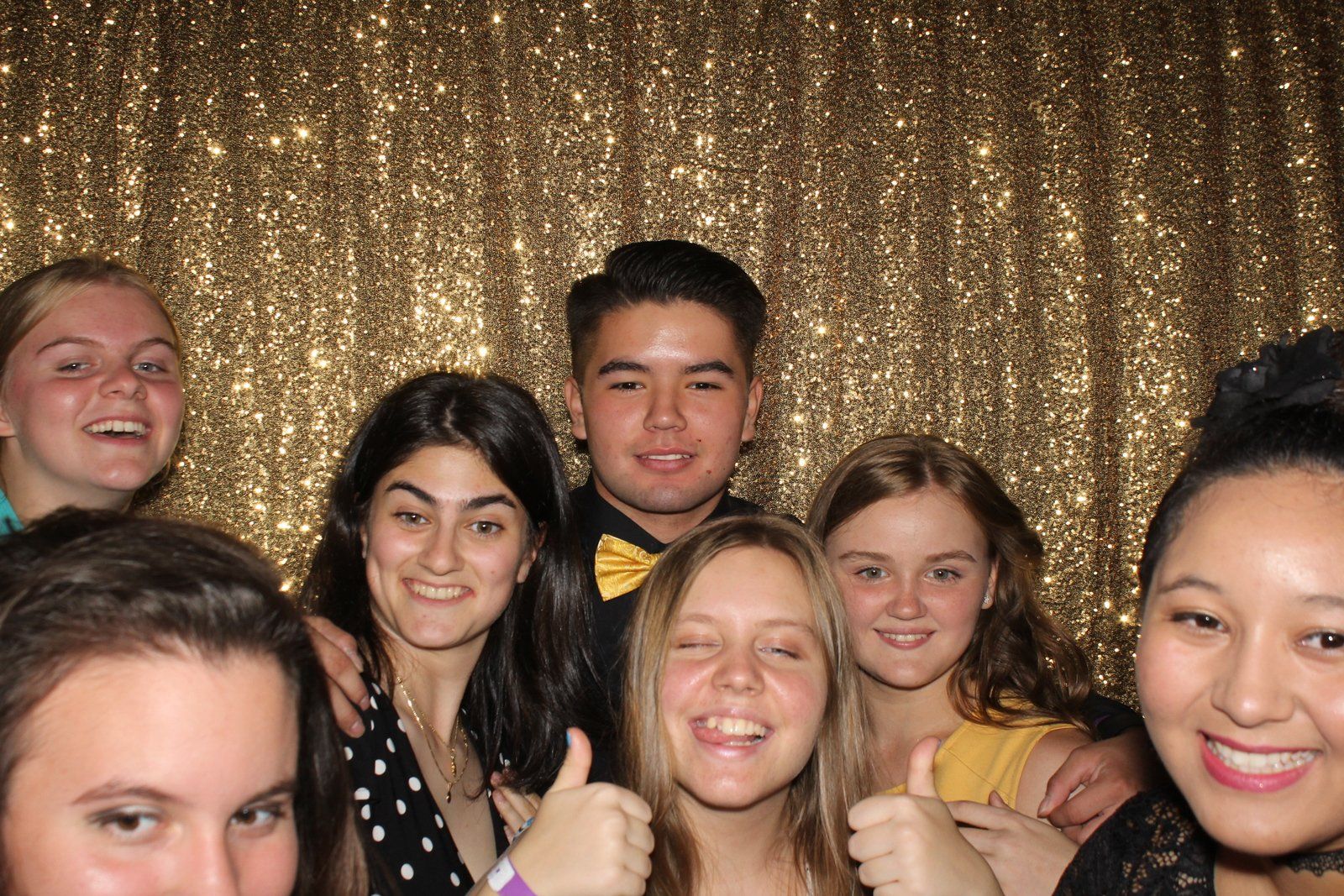 Group of people smiling and posing in front of a sparkly gold backdrop. Thumbs up.