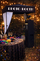 Black photo booth with a white backdrop, sitting on a yellow surface. A silver screen is attached to the side.