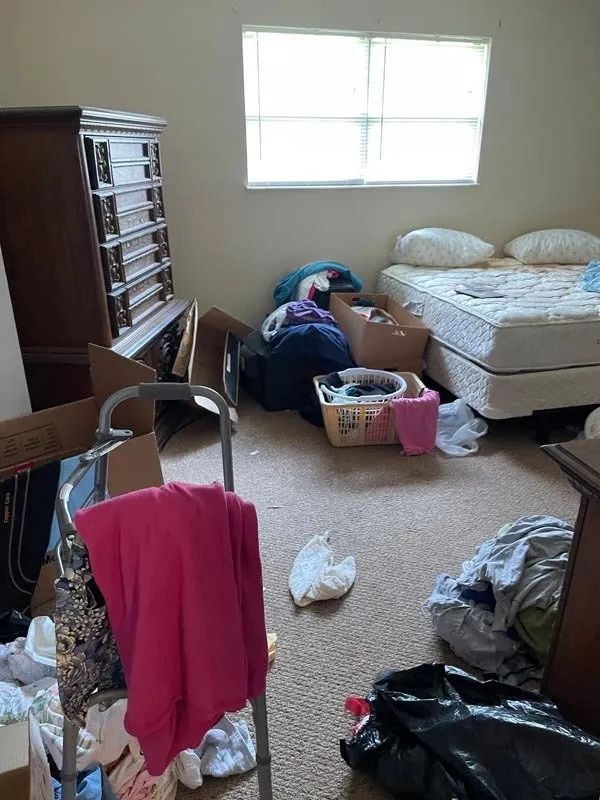 Cluttered bedroom with unmade bed, scattered clothes, laundry baskets, and a dresser by a bright window