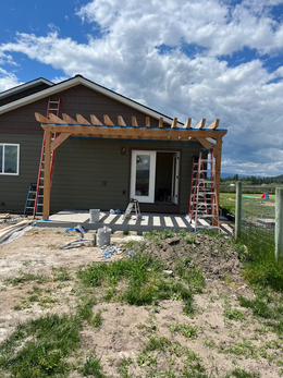 Small house exterior under construction with wooden pergola frame, ladders, and a partly cloudy sky.