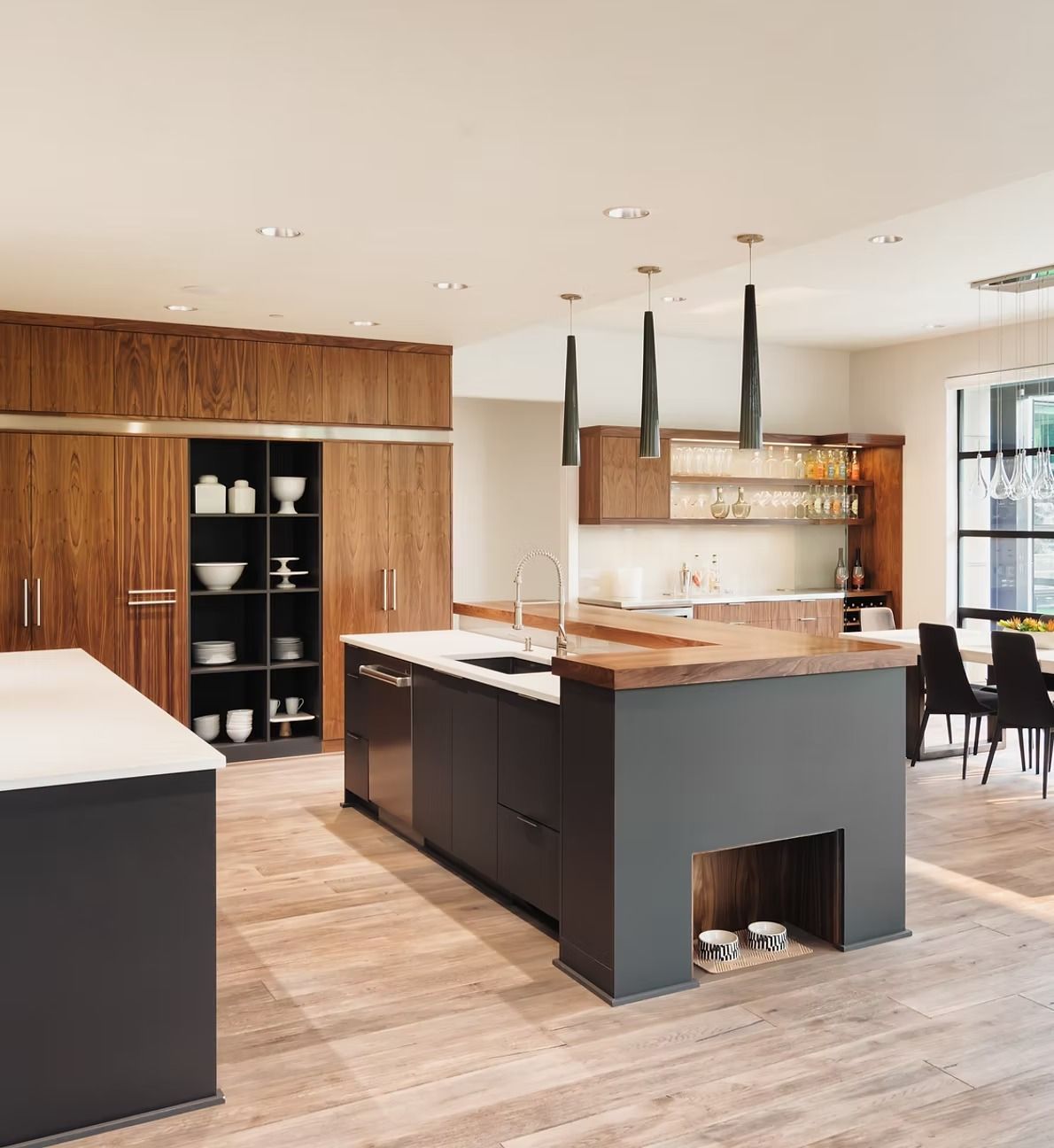 Modern kitchen with a large island, wood cabinetry, black accents, and pendant lights on a light wood floor.