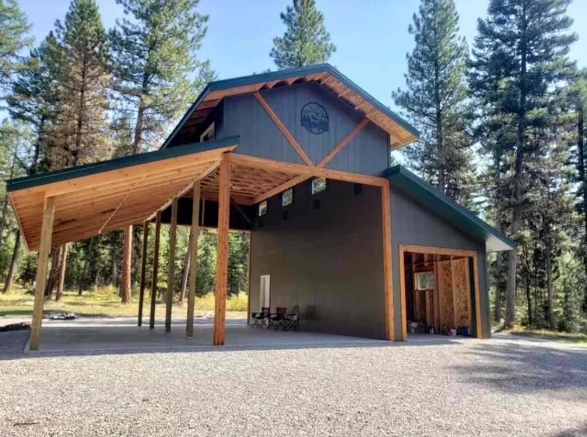 Modern cabin with a large wooden carport in a pine forest, on a gravel driveway.