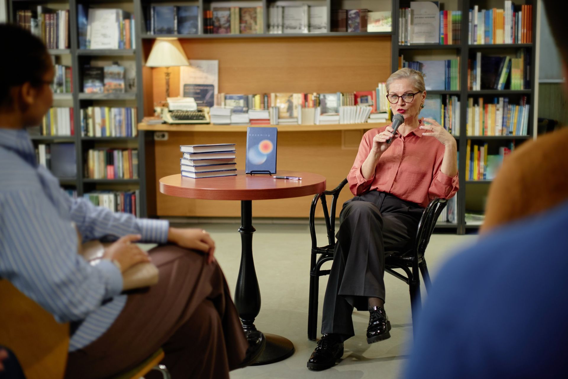 Two people seated in a library-like room, listening to a man speaking beside a round table and bookshelf.