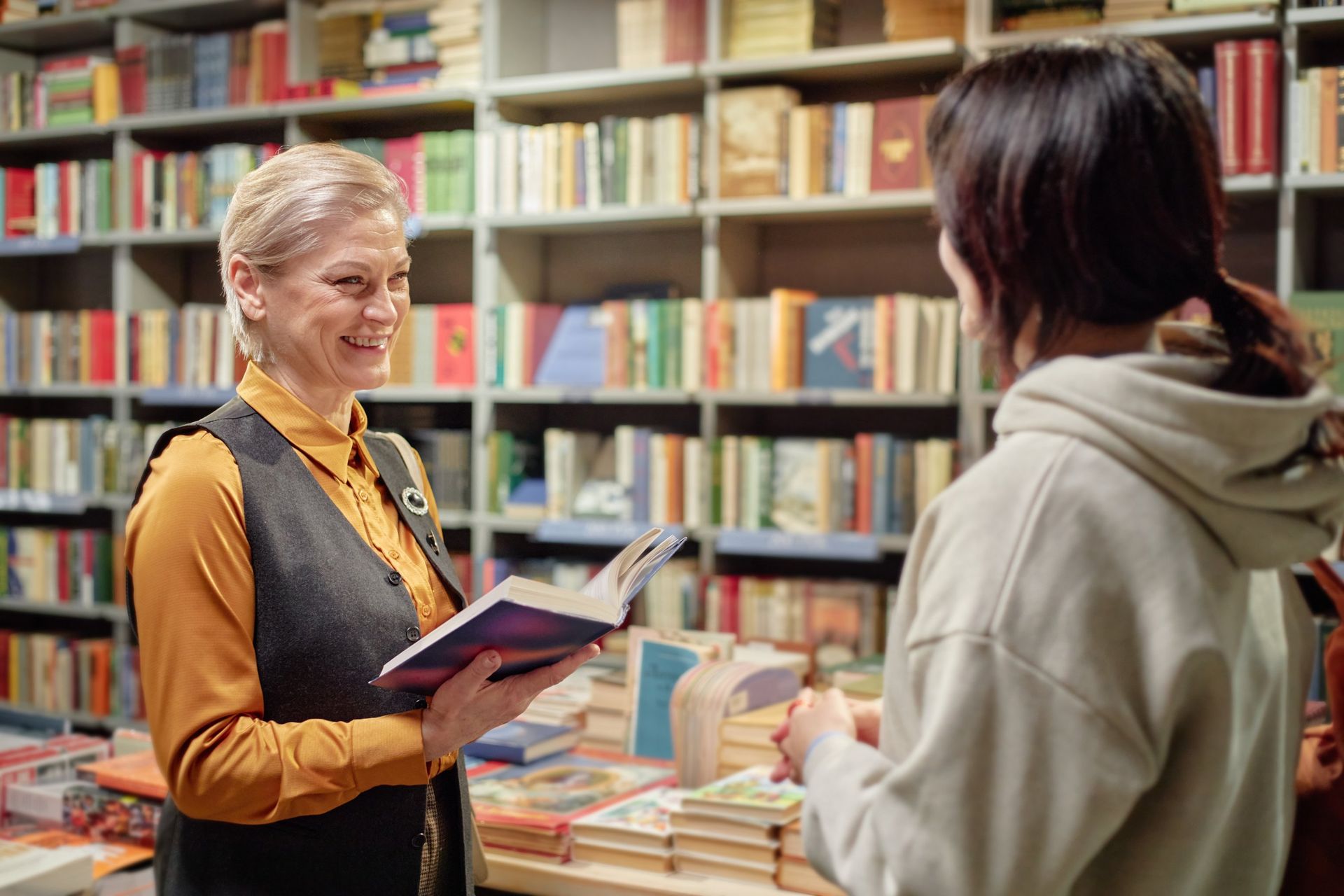 A smiling librarian in an orange shirt and vest holds an open book while speaking with a visitor in a bookstore aisle.