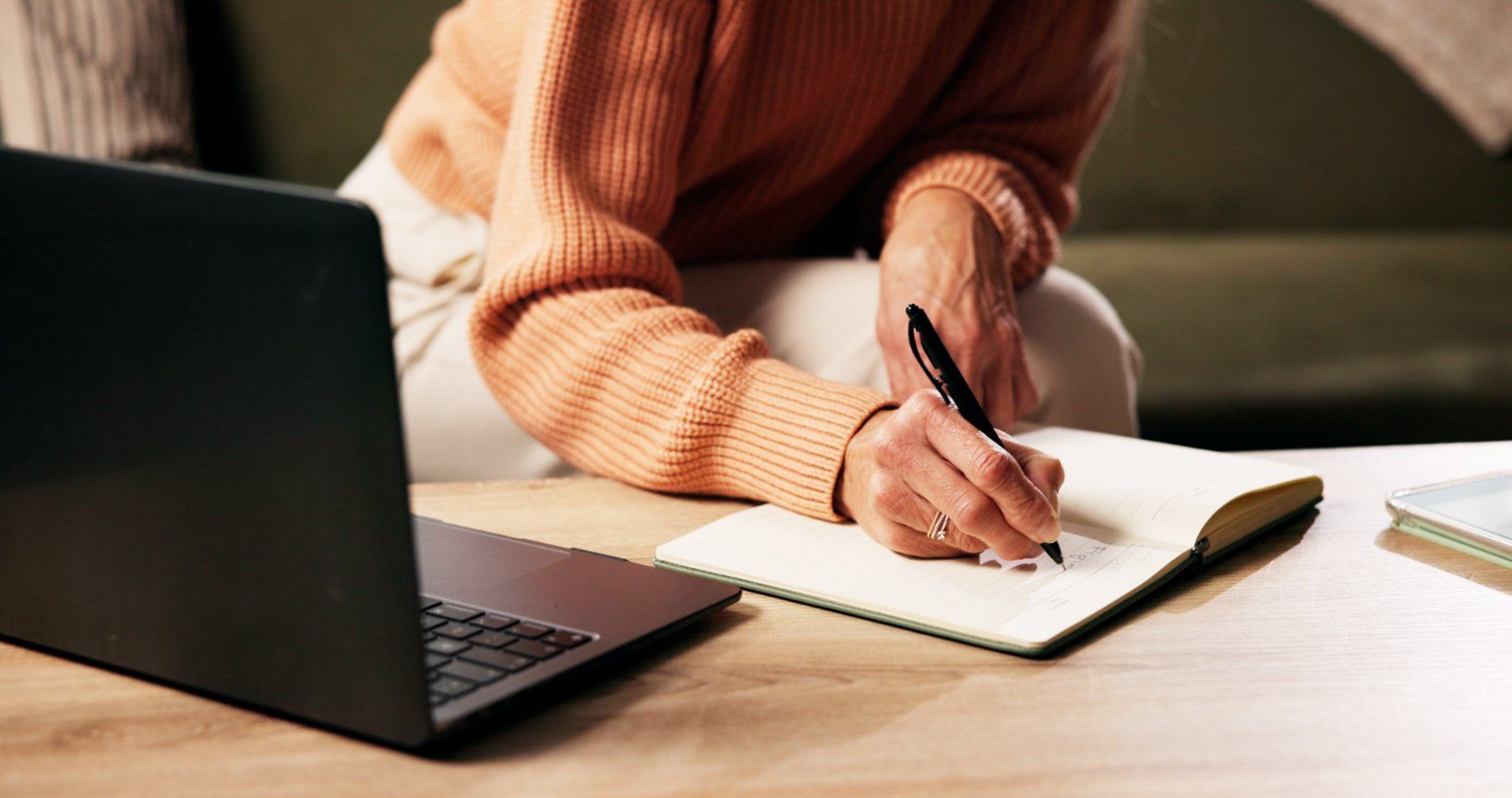 A person in a peach-colored sweater writes in a notebook while sitting at a table with a laptop.