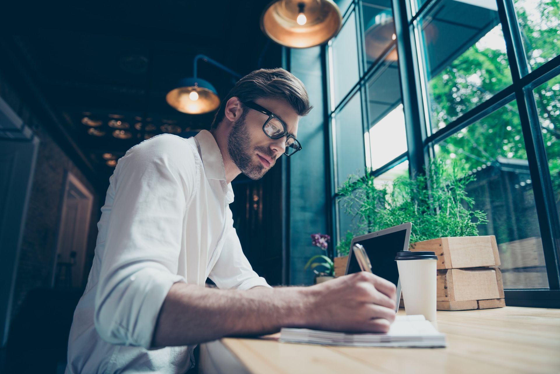 Man writing in a notebook at a sunlit café table by a window