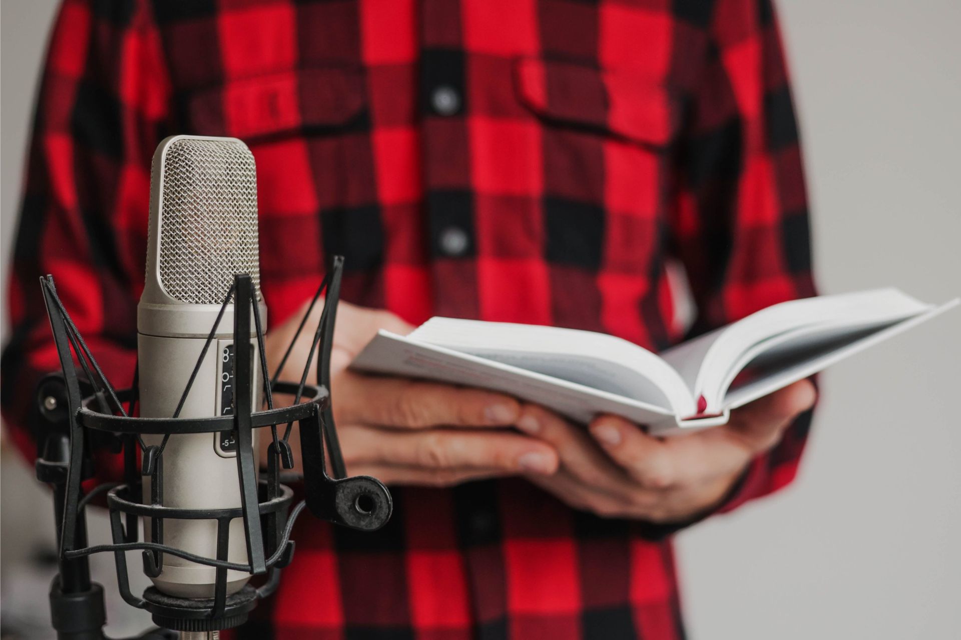 A person in a red and black plaid shirt holds an open book while standing in front of a studio microphone.