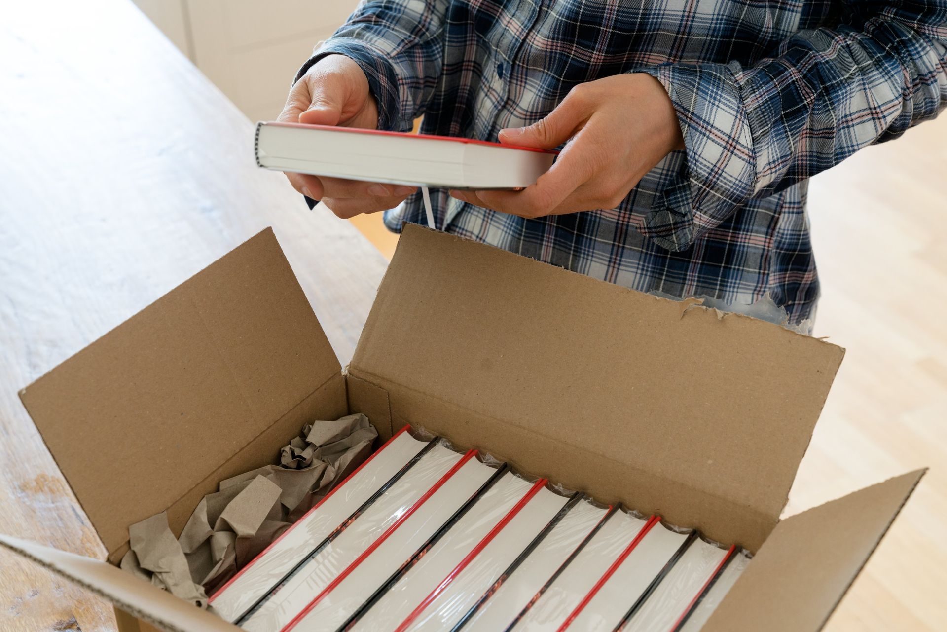 A person in a plaid shirt carefully places a book into an open cardboard box filled with several neatly packed books.
