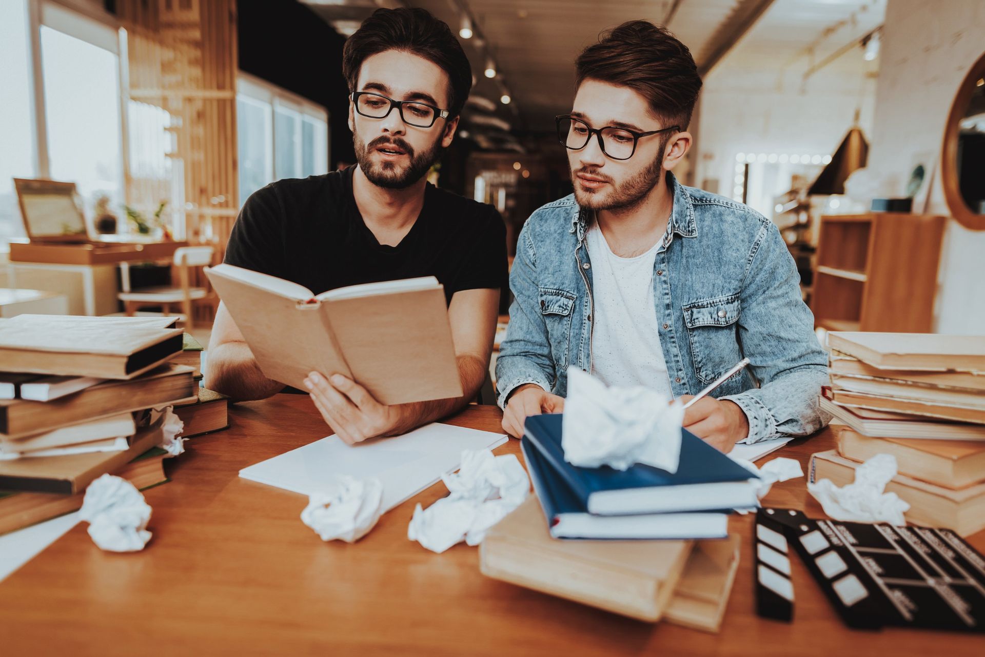 Two people with glasses sit at a cluttered desk reading a book, surrounded by stacks of papers and crumpled paper balls.