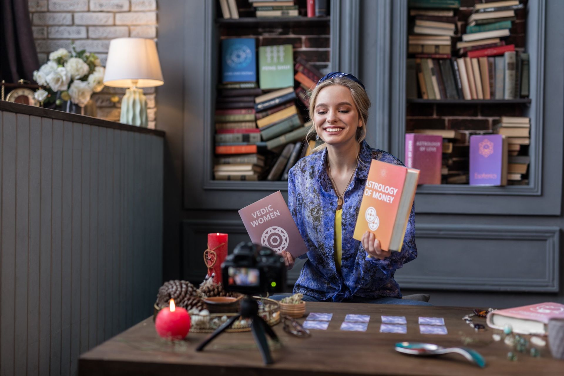 Woman smiling at a desk holding two books in a cozy book-lined room