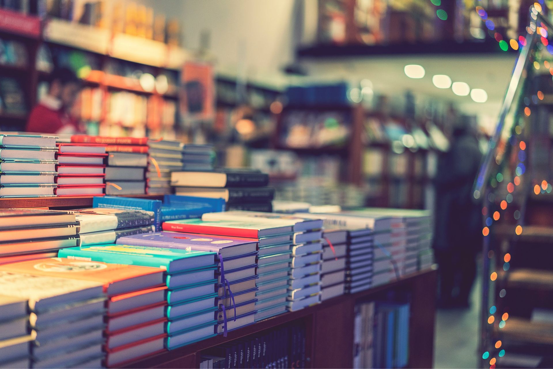 Stacks of colorful books on a wooden display table inside a softly lit, out-of-focus bookstore.